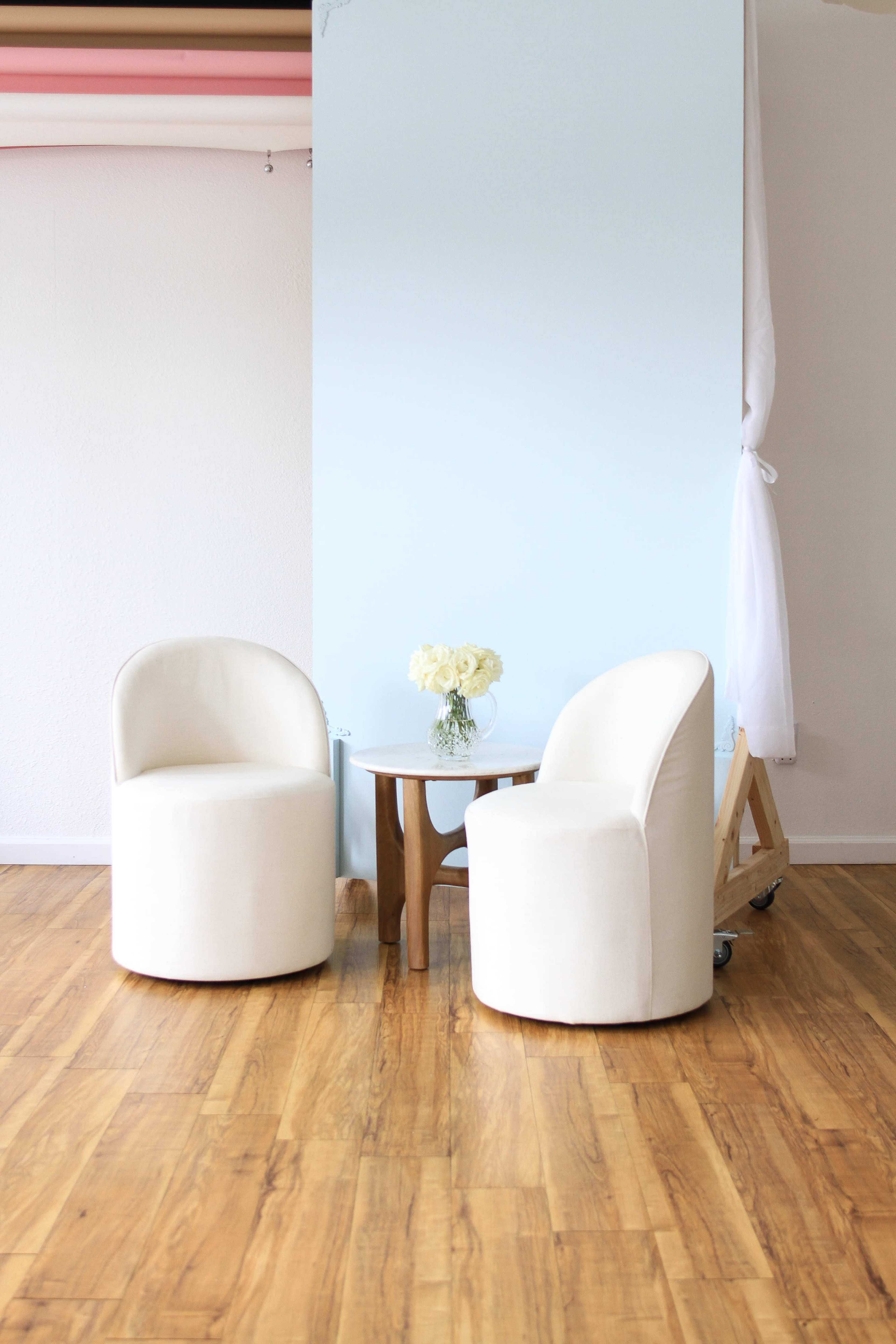 The image shows two cream-colored chairs positioned near a small wooden table with a vase of white flowers, set against a light blue wall in a room with wooden flooring.