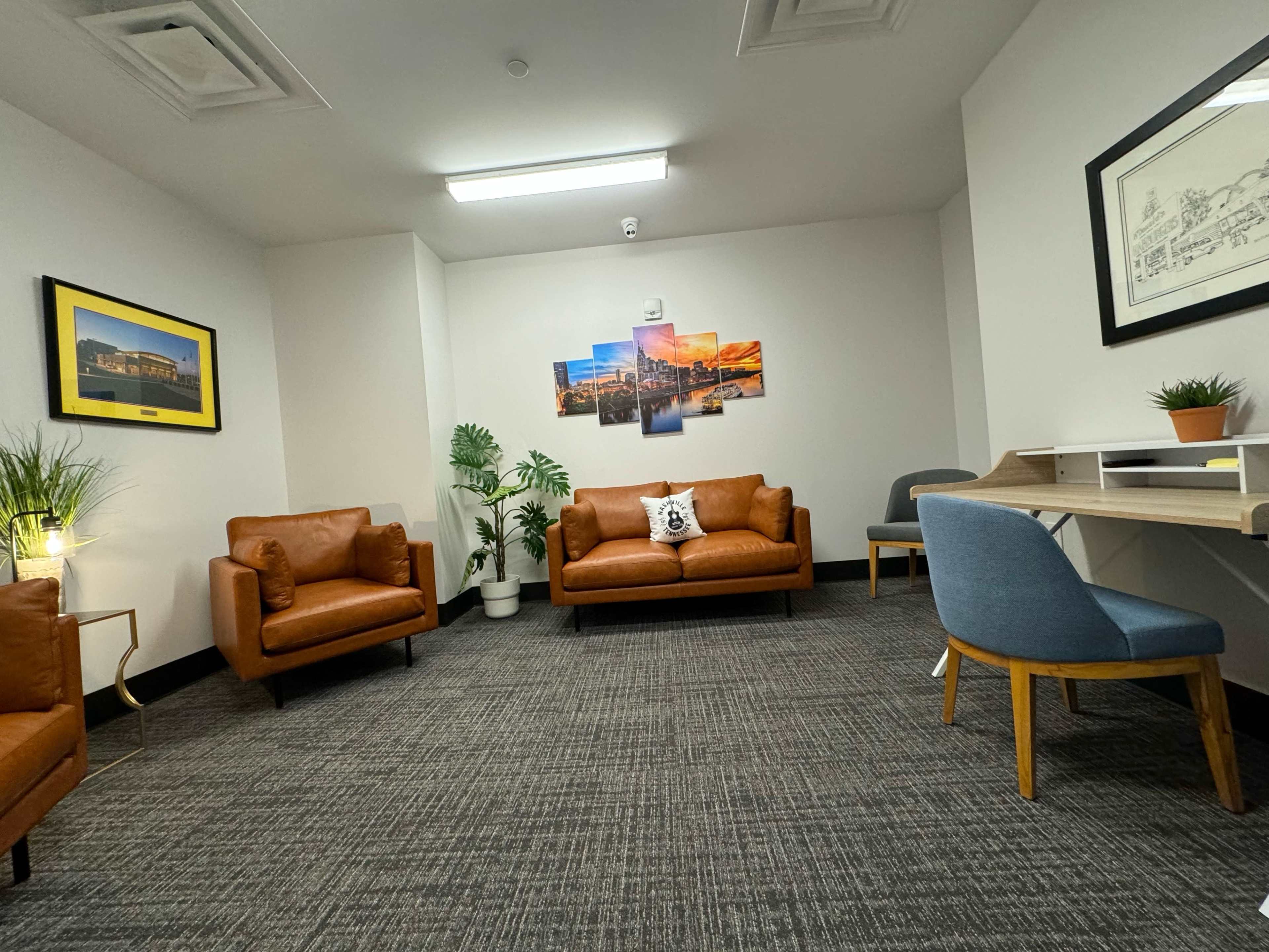 The room features two brown leather sofas, a blue chair, a desk, and decorative plants, with artwork on the walls.
