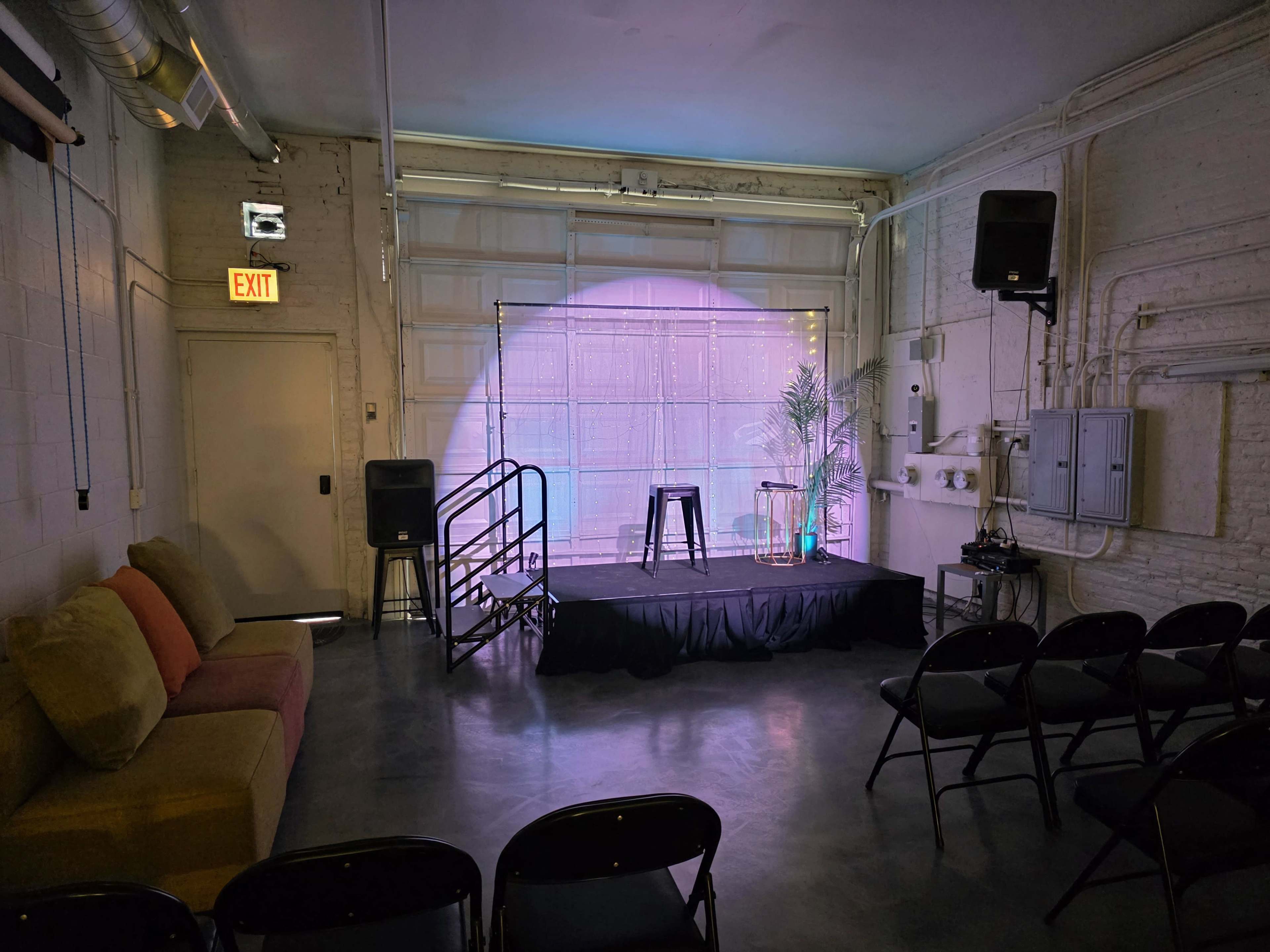 The image shows a small performance space with a stage featuring a stool and a backdrop, arranged with rows of black chairs facing the stage and a couch to the side.