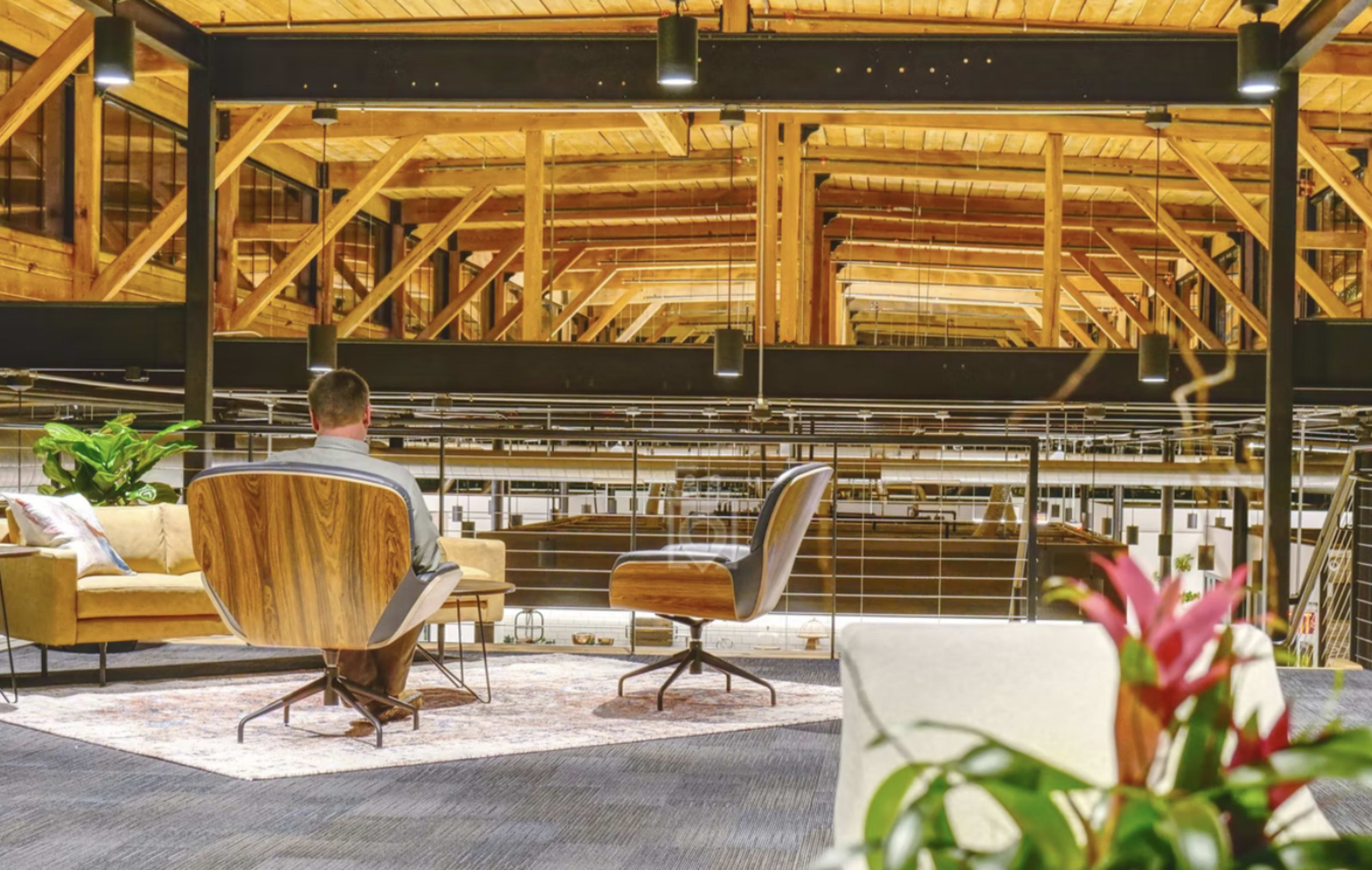 A person sits in a modern, open-concept space with wooden beams, overlooking a lower level furnished with seating and greenery.