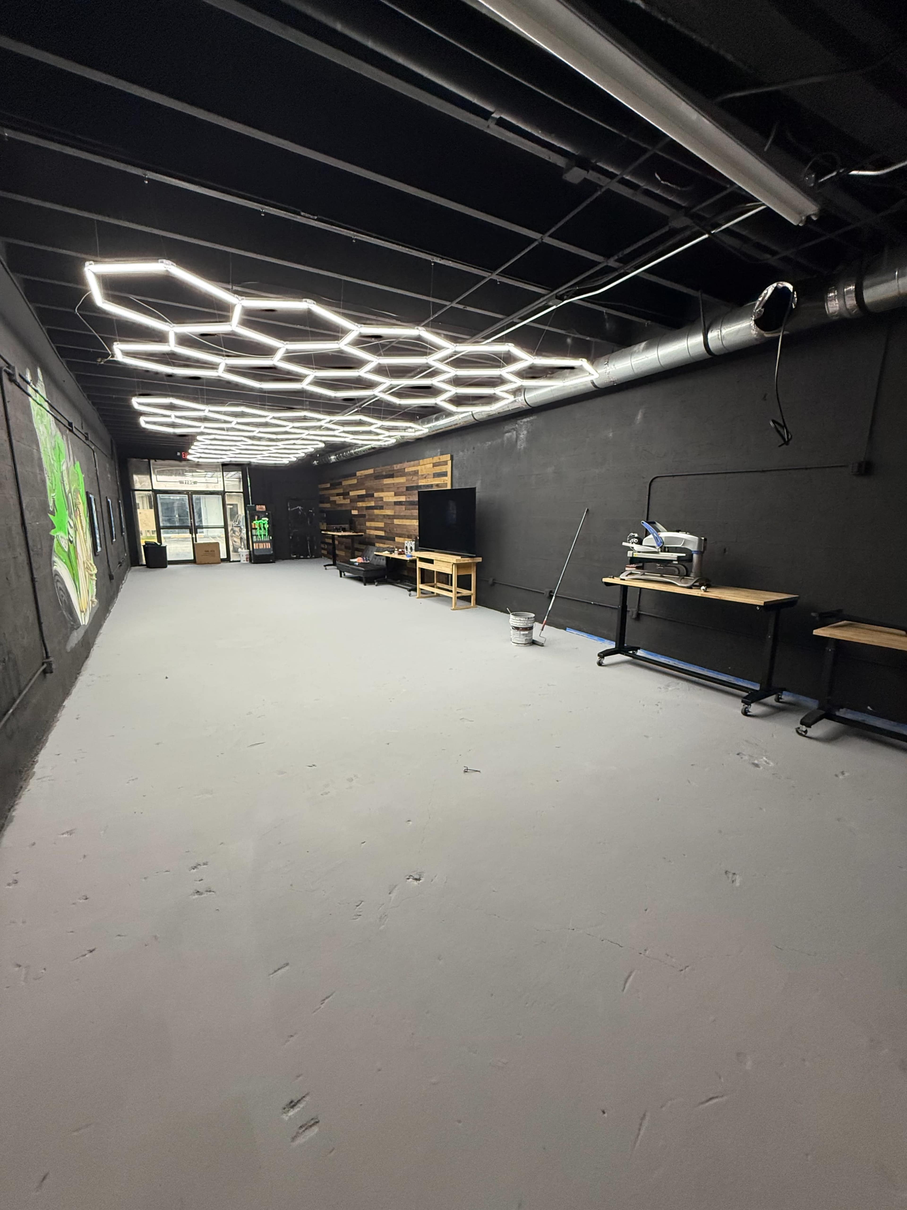 The image shows an empty indoor space featuring hexagonal ceiling lights, a television, and work tables against a black wall.