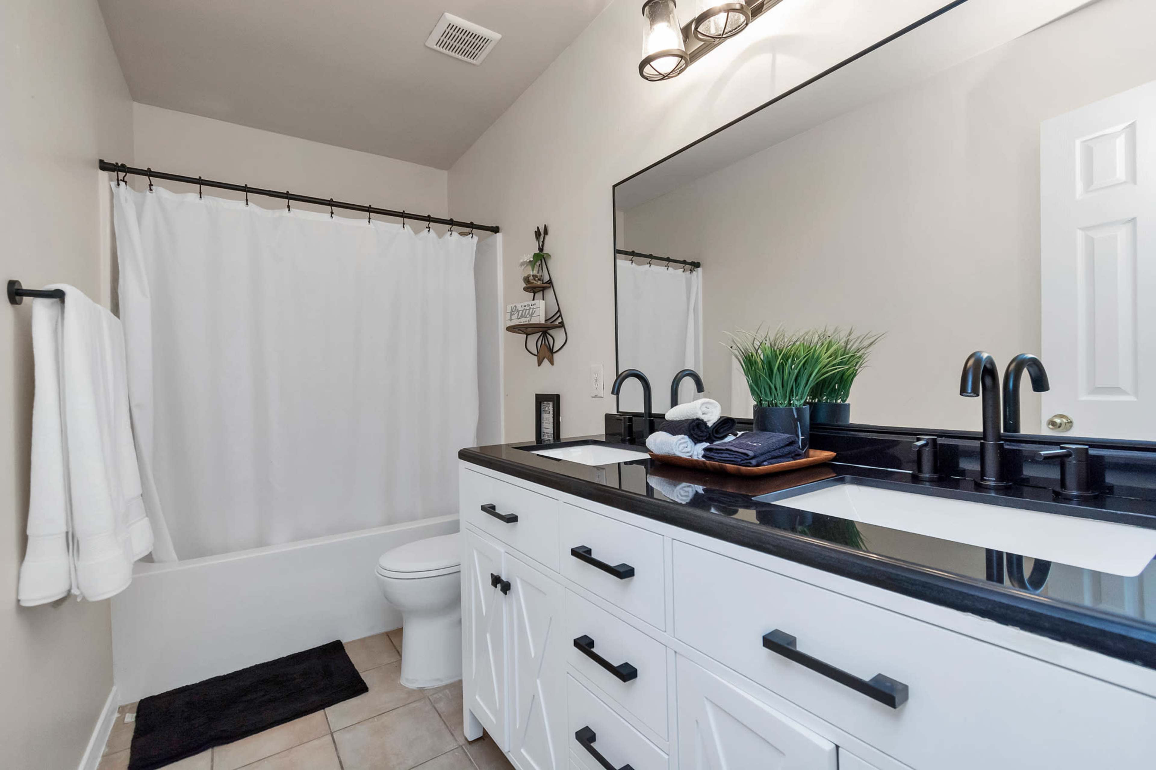 A modern bathroom features a white shower curtain, a black countertop with two sinks, and a large mirror above a white cabinet.