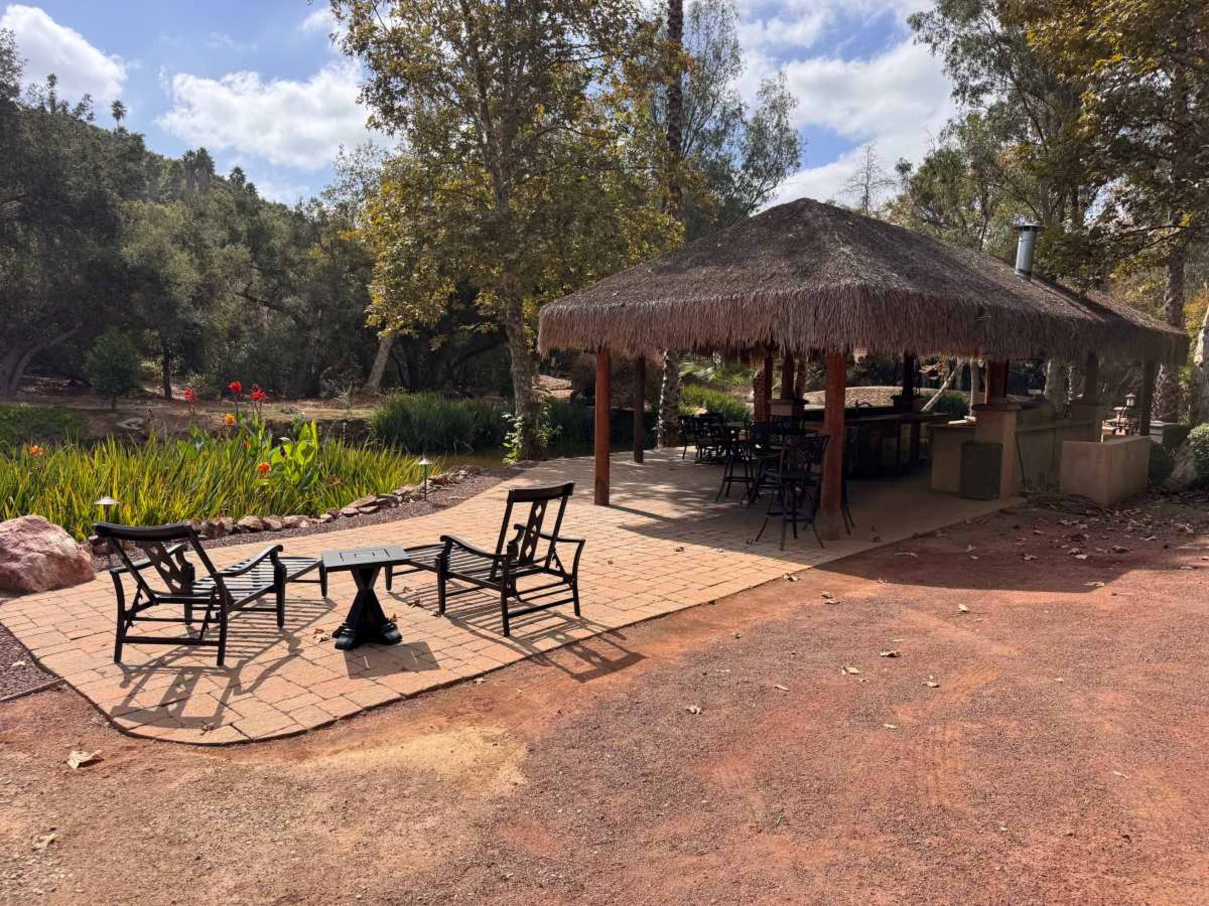 A thatched-roof pavilion with seating and a stone patio is surrounded by greenery and trees.