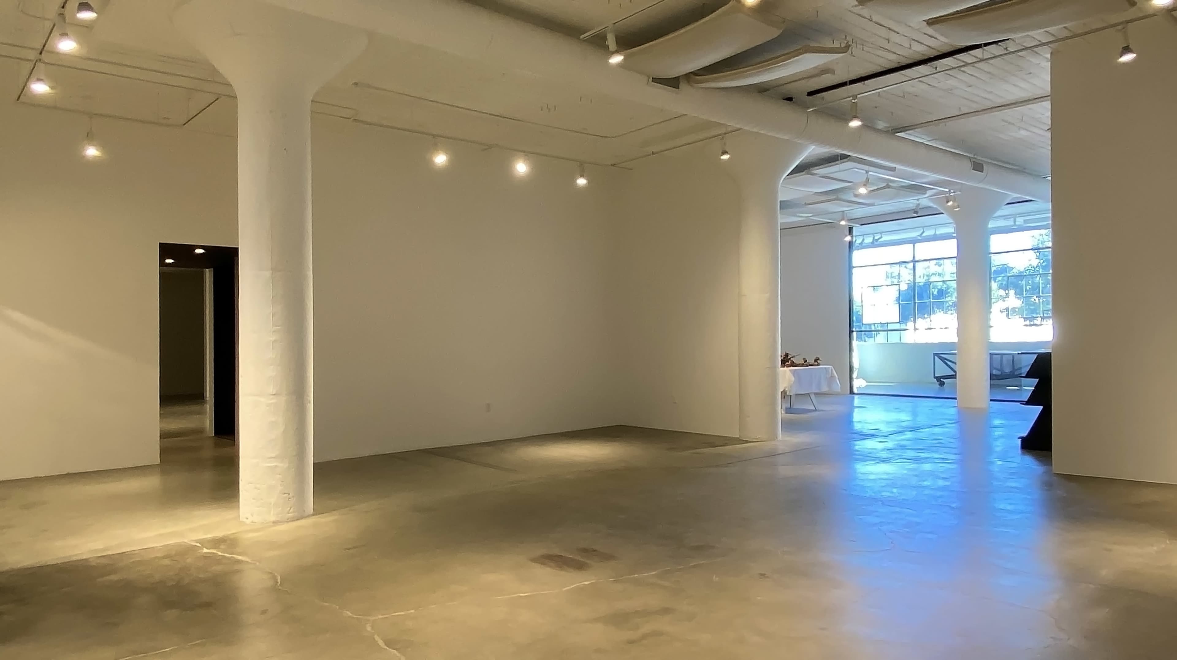 The image shows a spacious, well-lit interior of an art gallery with concrete flooring and large windows.