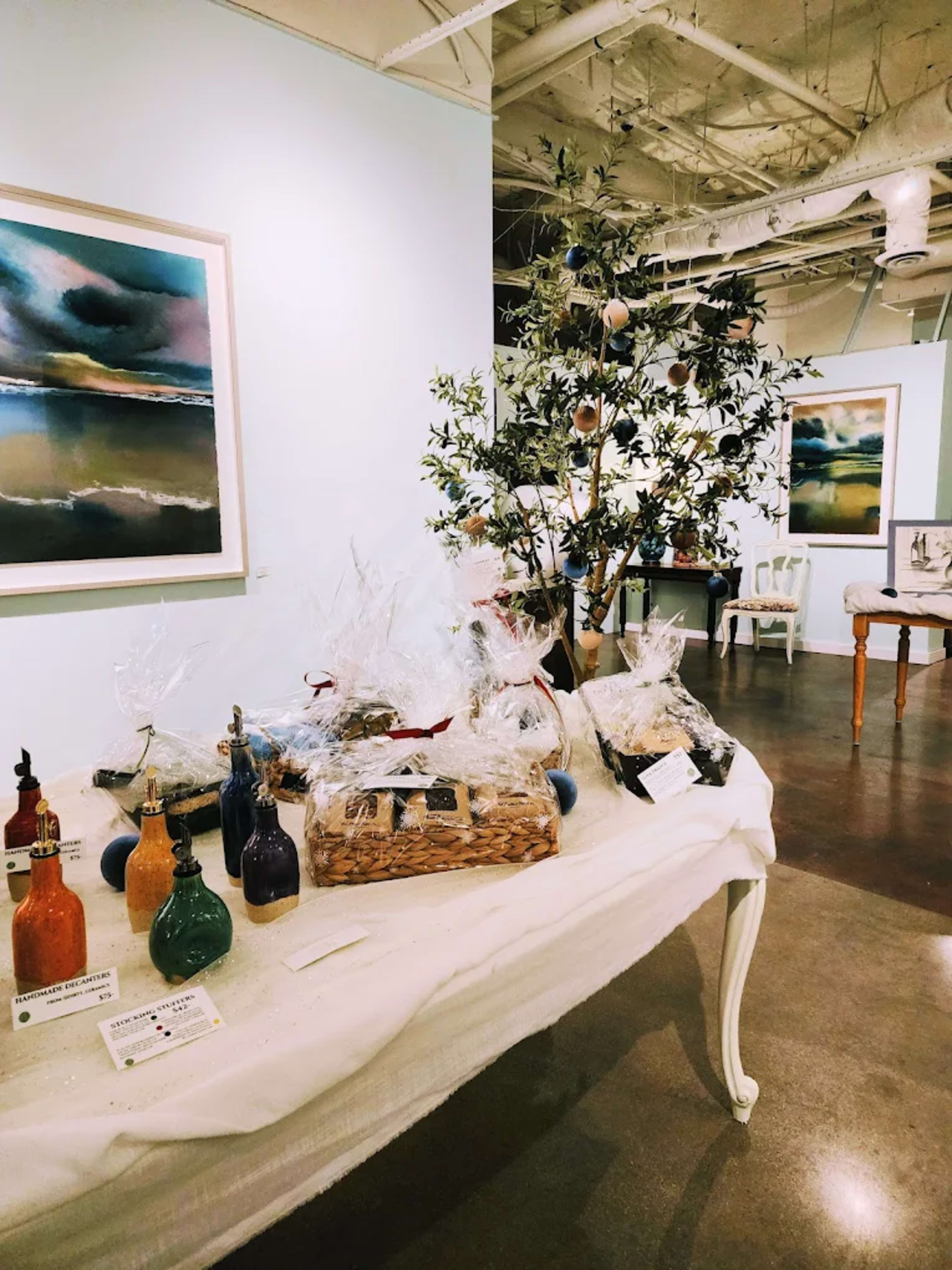 The image shows a gallery interior featuring a table with various decorative bottles and gift baskets, alongside a small tree adorned with ornaments and artwork displayed on the walls.