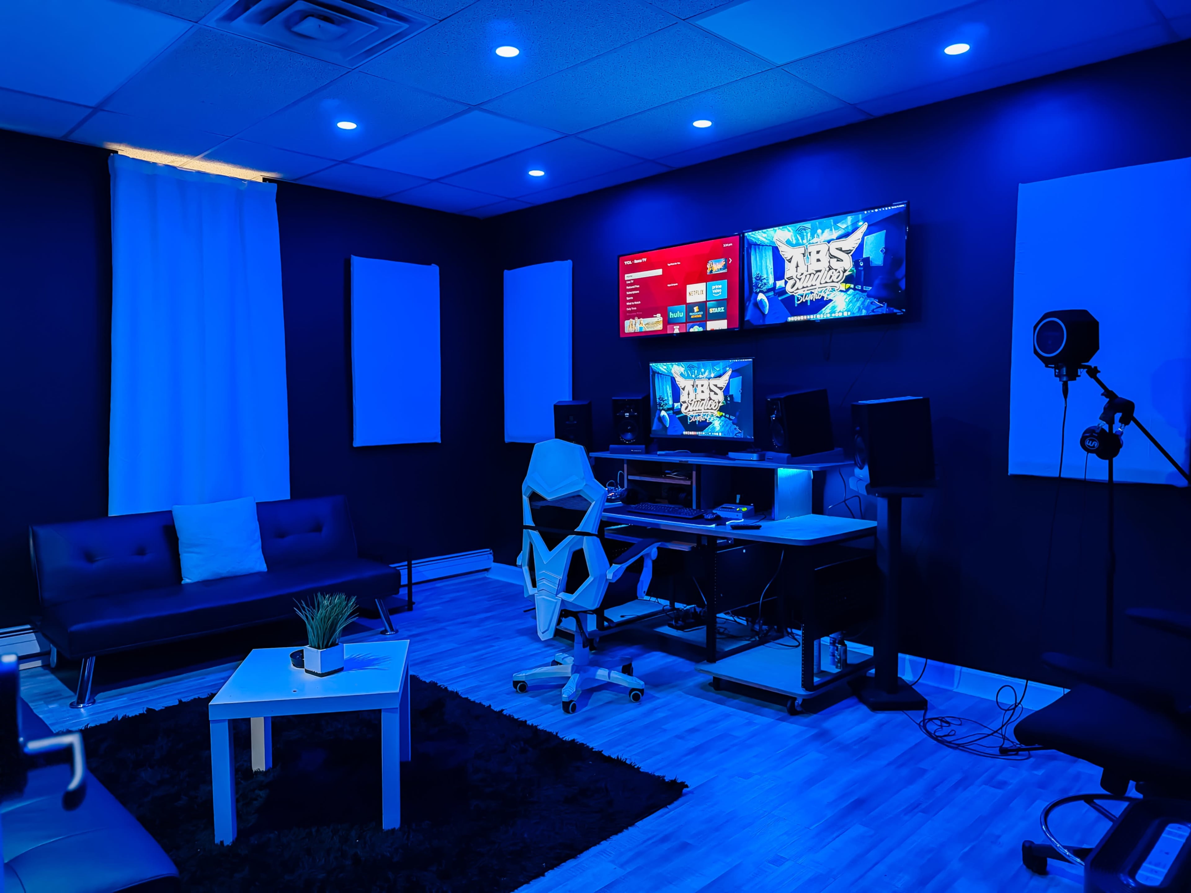 The image displays a modern recording studio featuring a black and blue color scheme, with a couch, a white coffee table, computers, and audio equipment set against the walls.