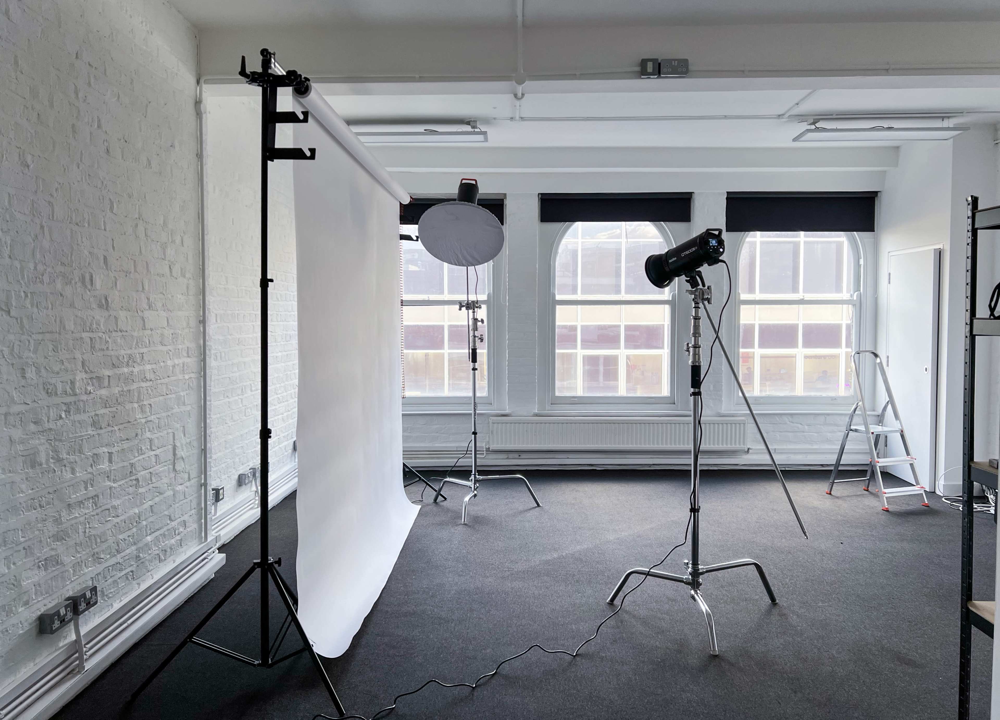 A photography studio features a white backdrop, lighting equipment, and a ladder in a spacious room with large windows.