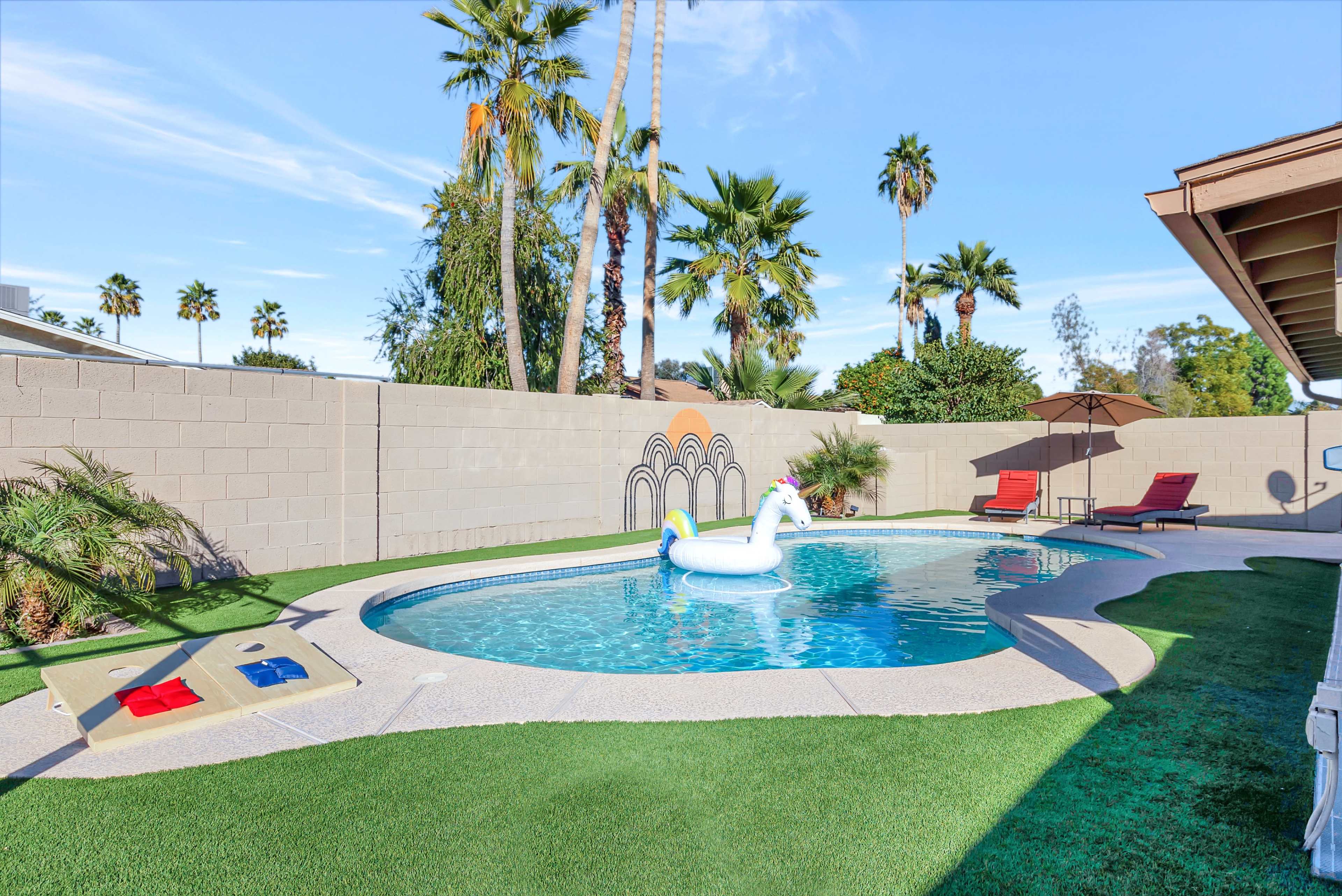 A backyard swimming pool with a unicorn float, surrounded by palm trees and two lounge chairs under a shade umbrella.