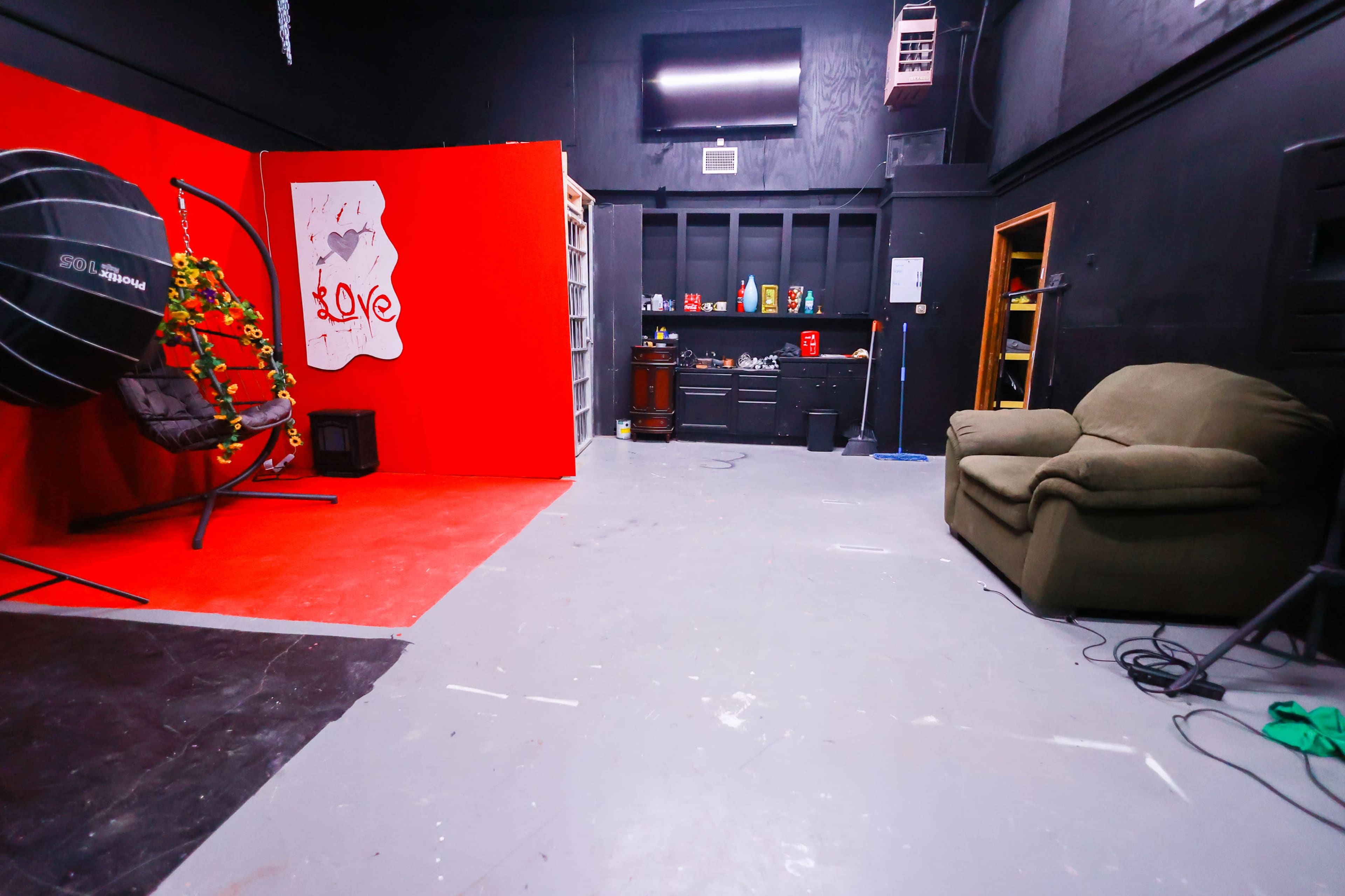 A photography studio features a red accent wall, a swing chair with decorations, shelves filled with various items, and a green couch in a sparse layout.