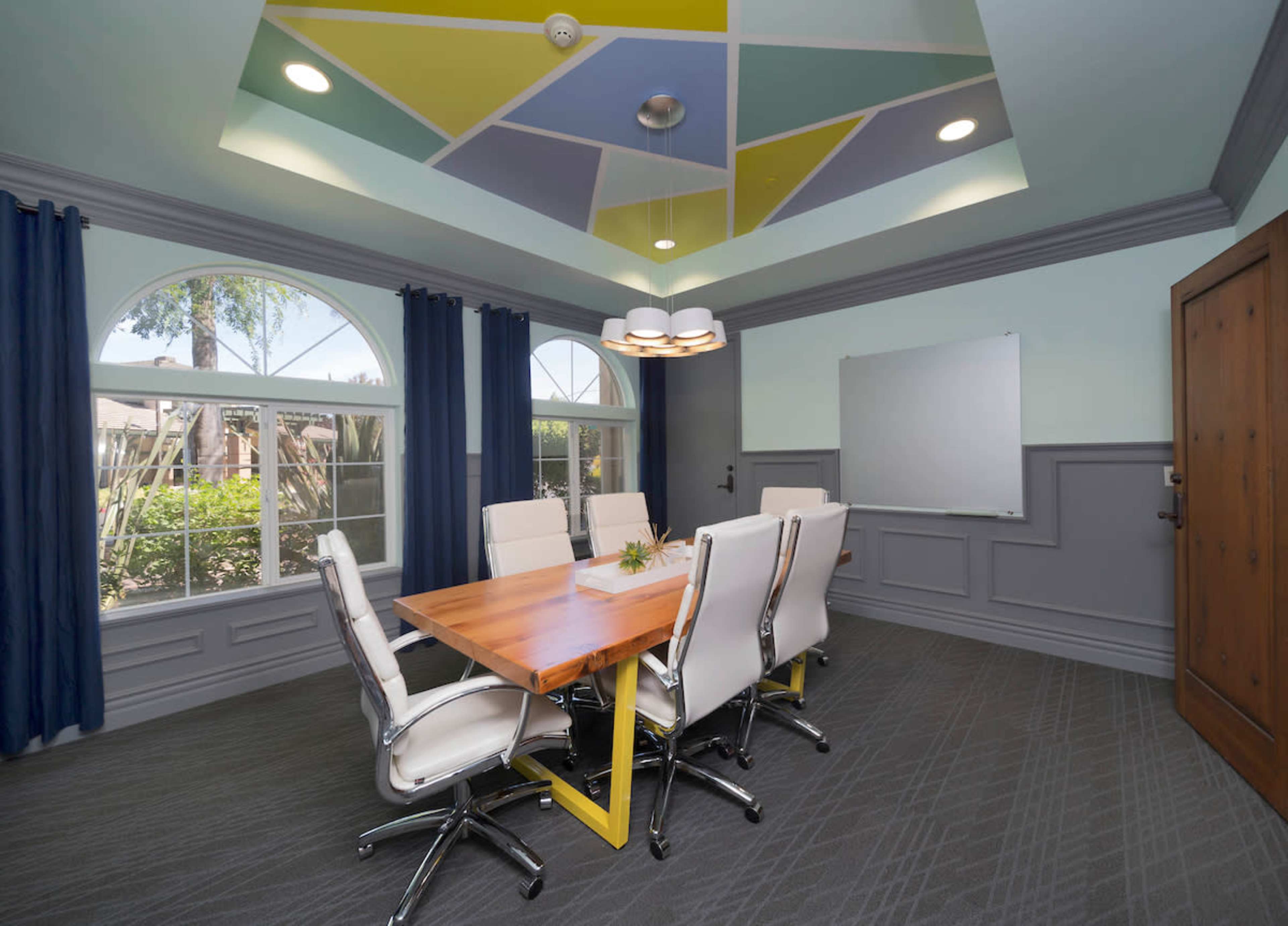 A modern conference room features a wooden table surrounded by white chairs, large windows, and a colorful geometric ceiling.