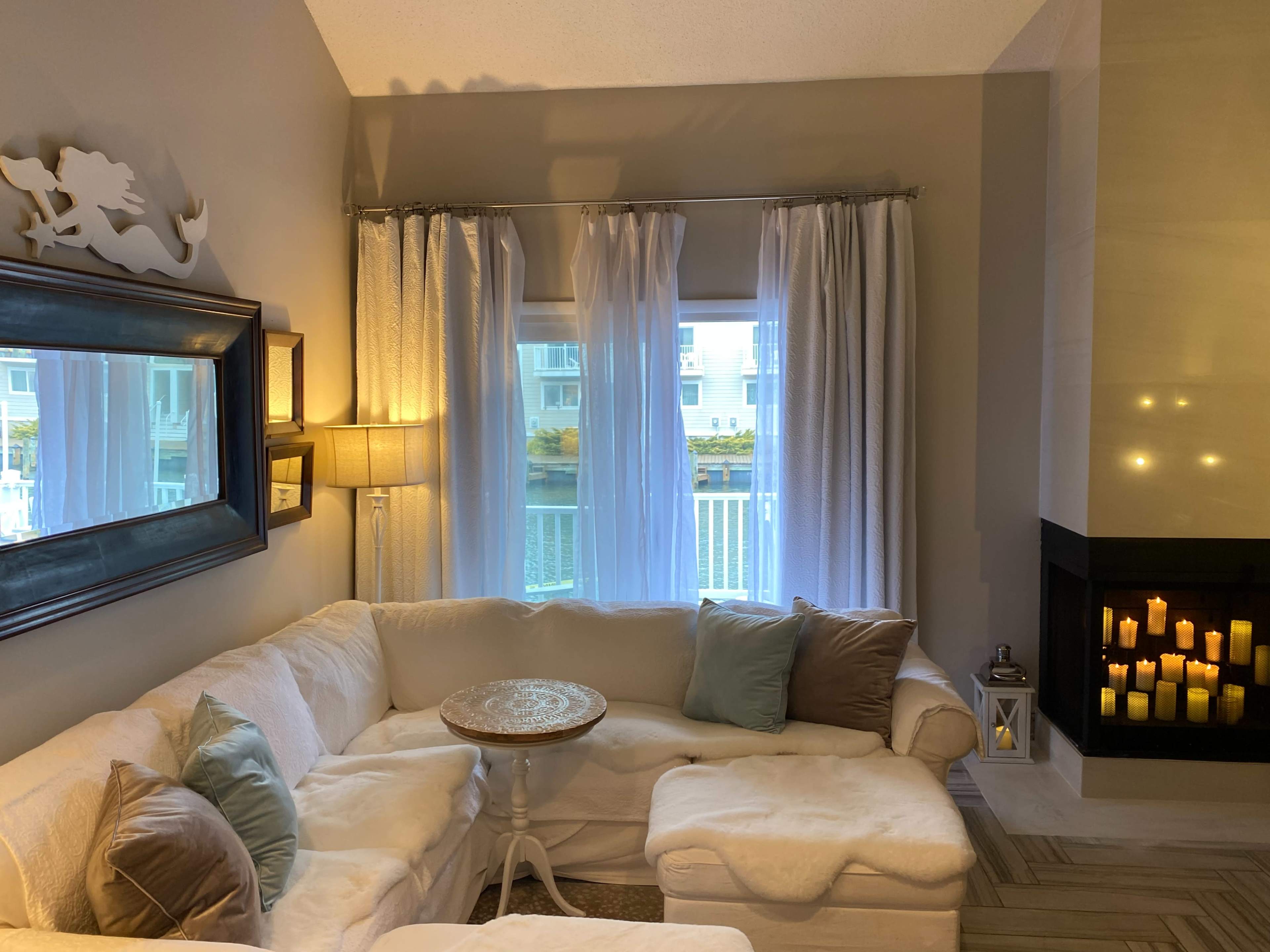 A cozy living room with a white sectional sofa, a small round table, and a fireplace illuminated by candles, all framed by sheer curtains and soft lighting.