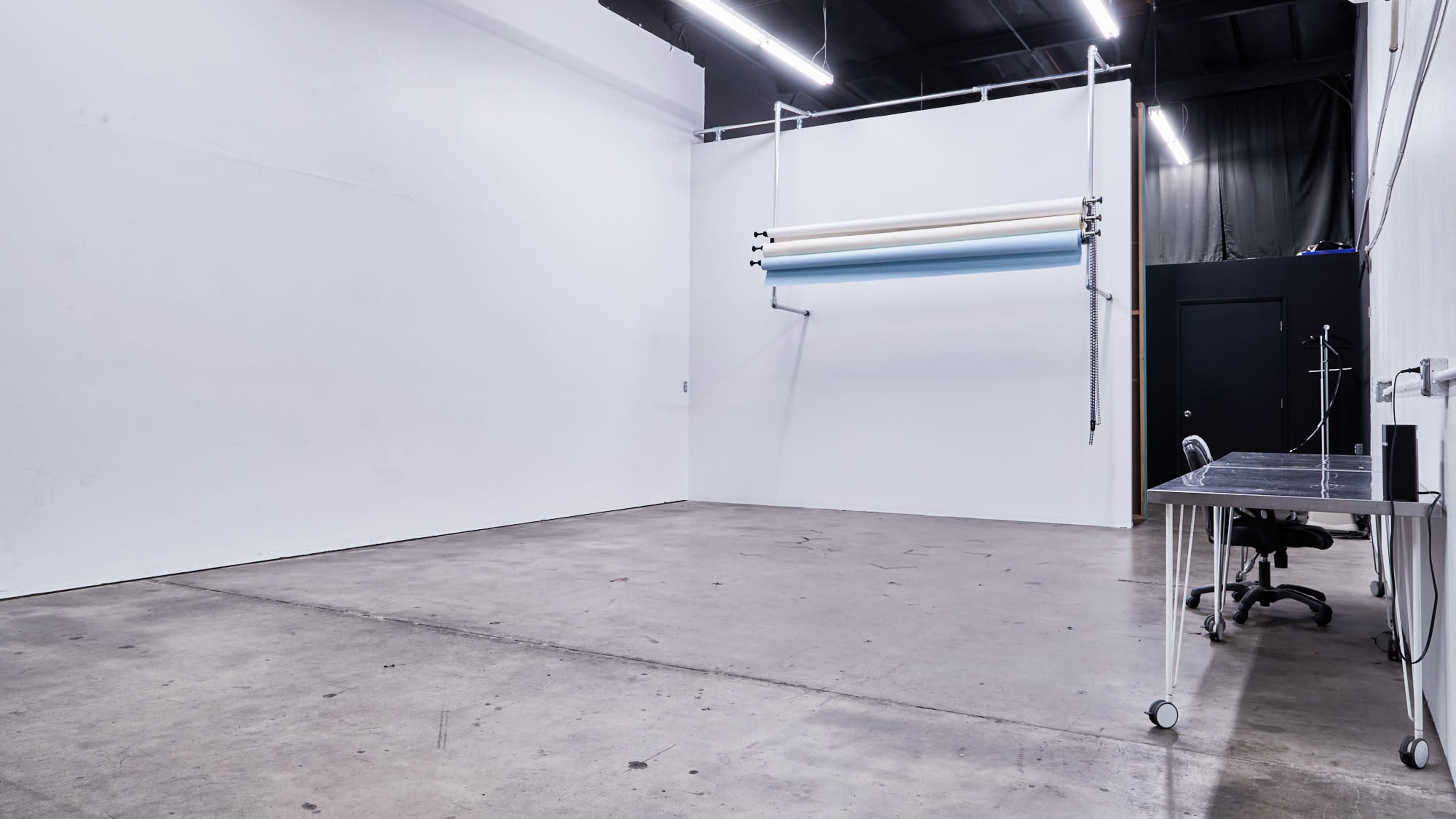 A spacious, empty studio with white walls, a backdrop system on one wall, and a metal table with a chair in one corner.