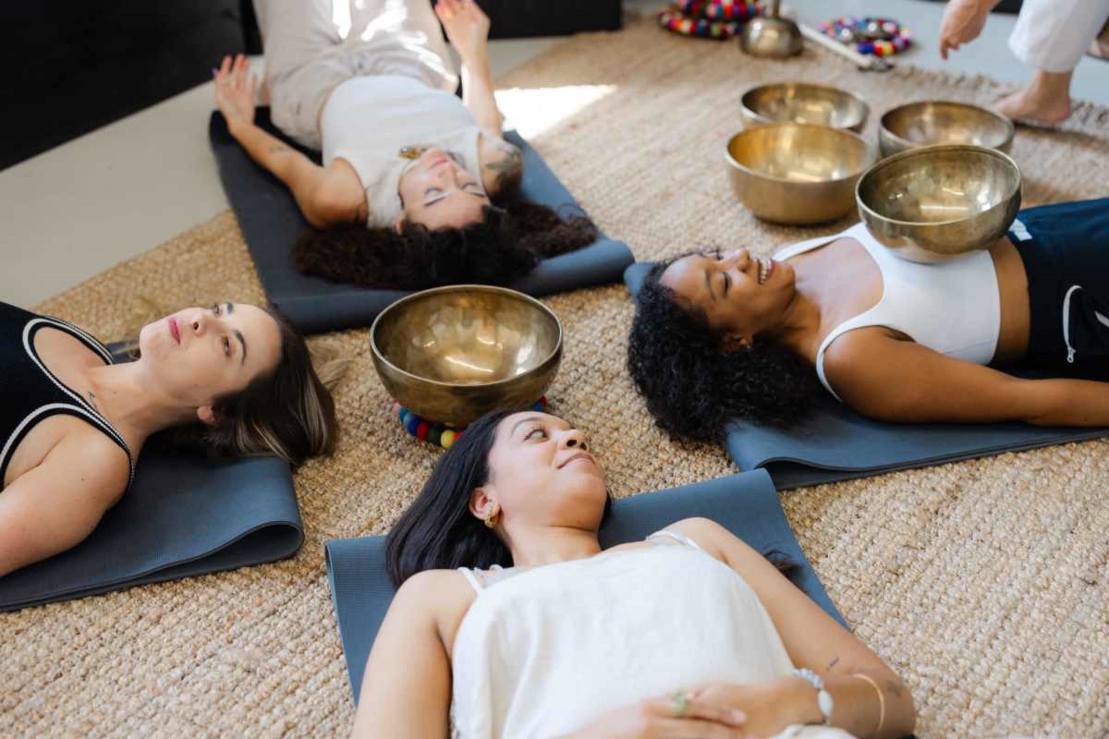 Four women are lying on yoga mats in a peaceful setting with several bowls placed on their chests.