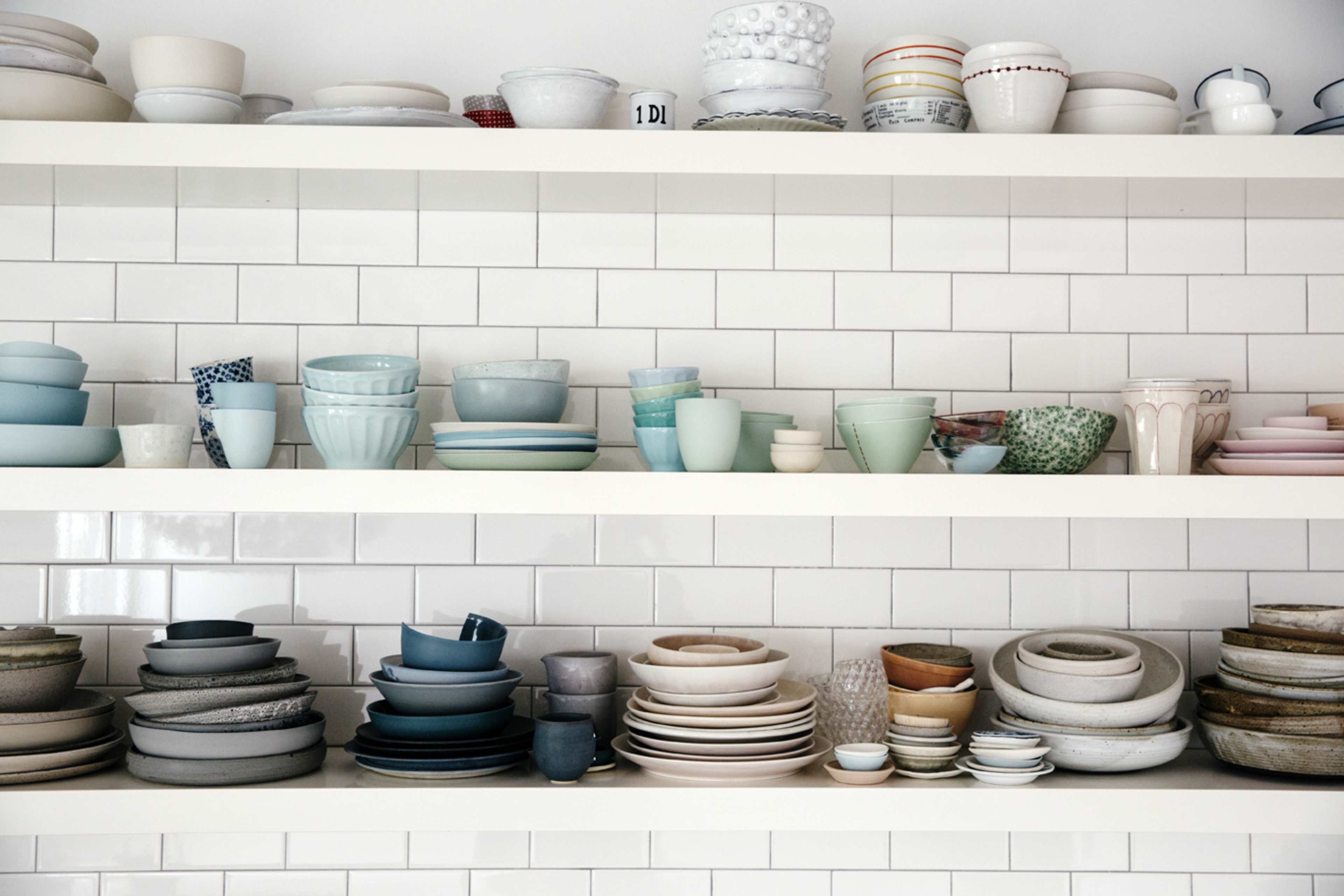 A collection of various plates, bowls, and cups is displayed neatly on two shelves against a tiled wall.
