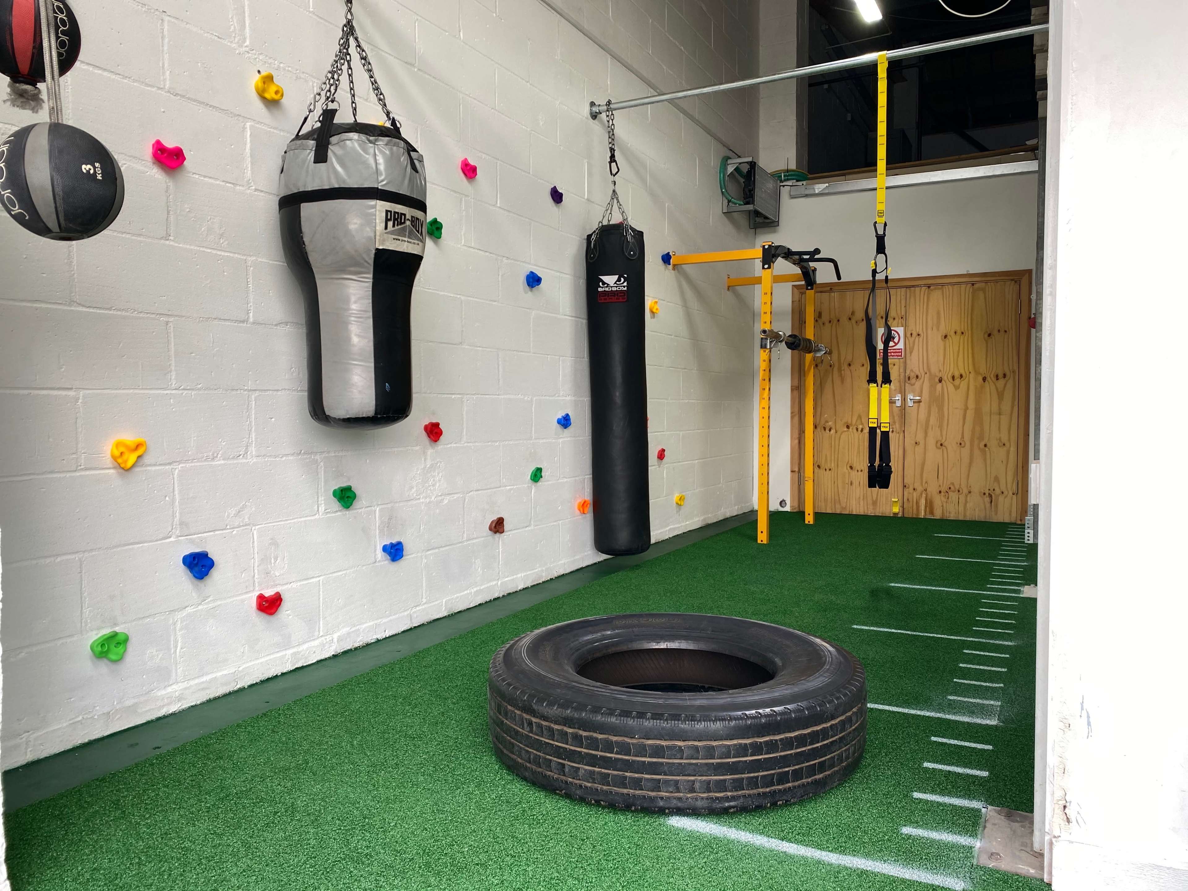 A small indoor workout space featuring a hanging punching bag, a battle rope setup, a tire on the green turf floor, and colorful climbing holds on the wall.