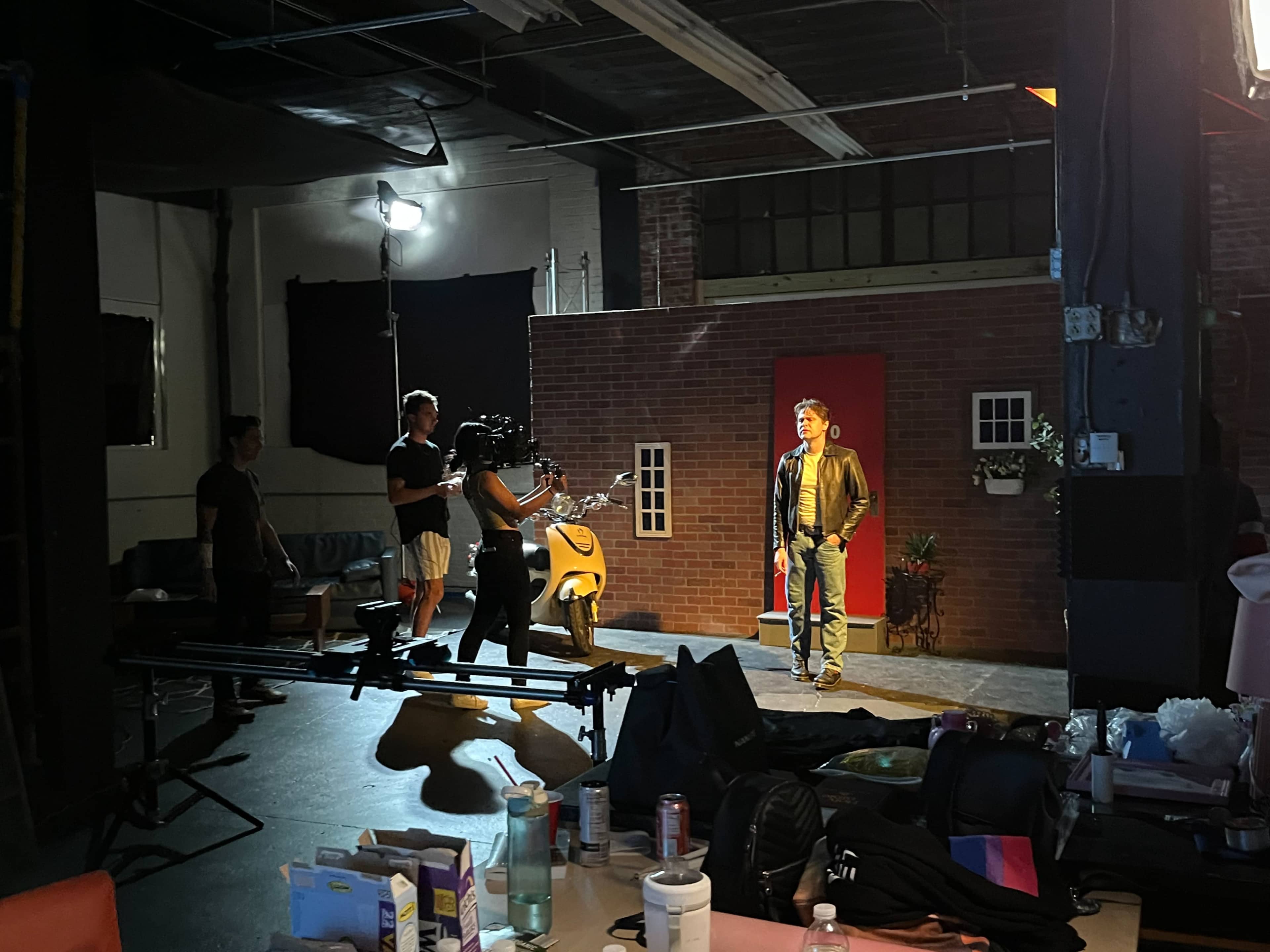 A film crew sets up in a studio, with a lead actor standing in front of a brick wall backdrop while a camera is positioned nearby.