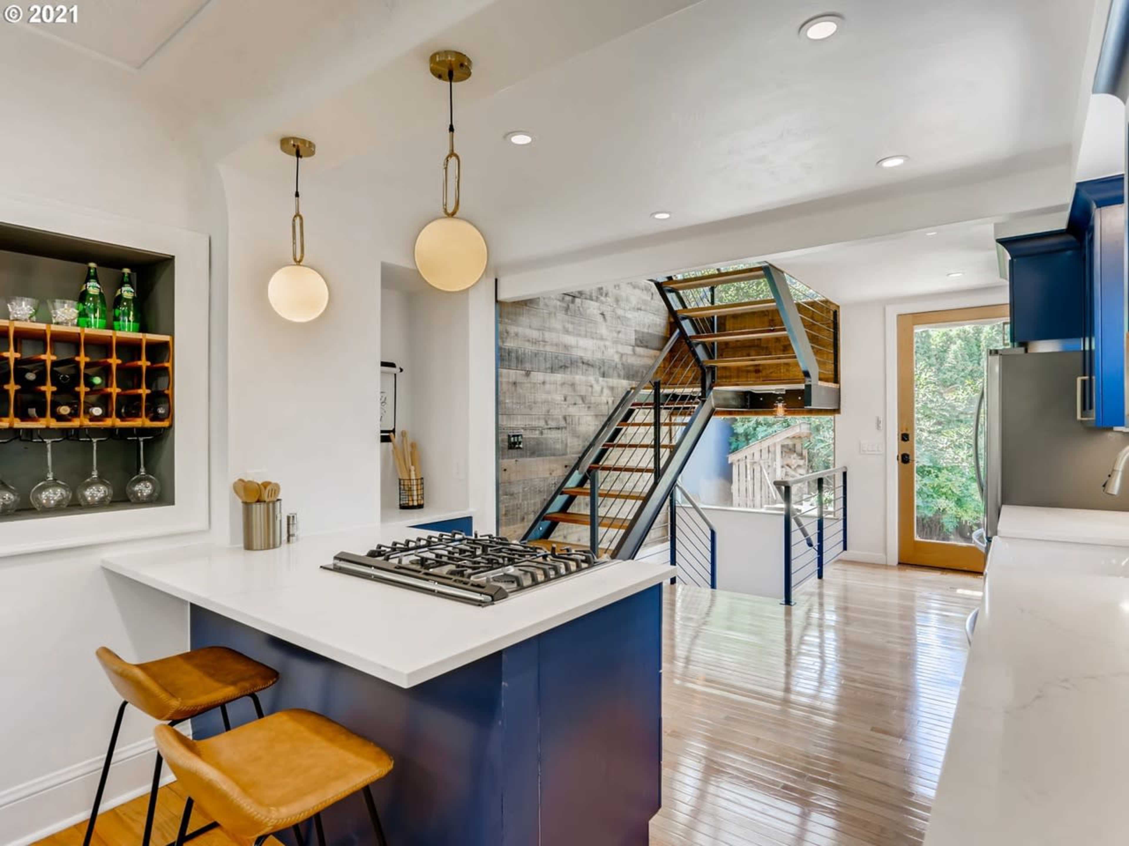 The image showcases a modern kitchen with a central island, bar stools, and a staircase leading to an upper level.