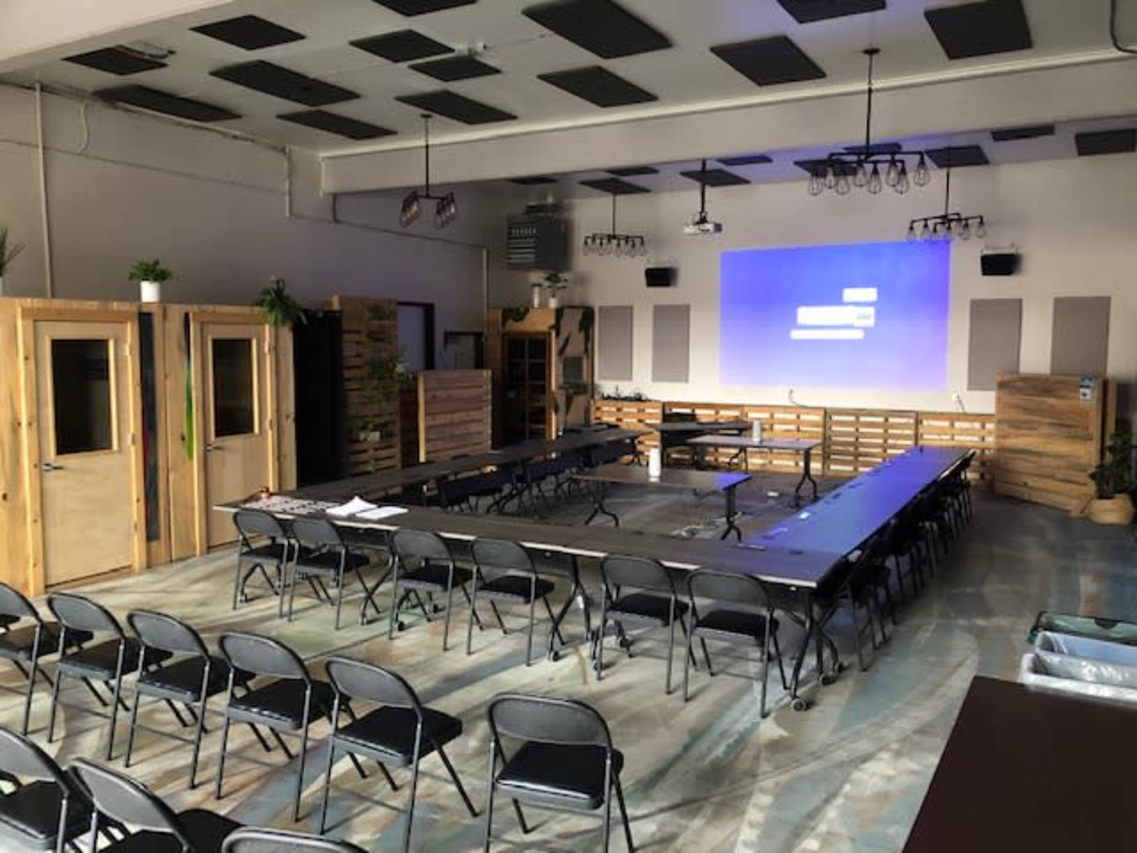 A spacious meeting room with black chairs arranged around a central table, and a projector screen displaying text on the wall.