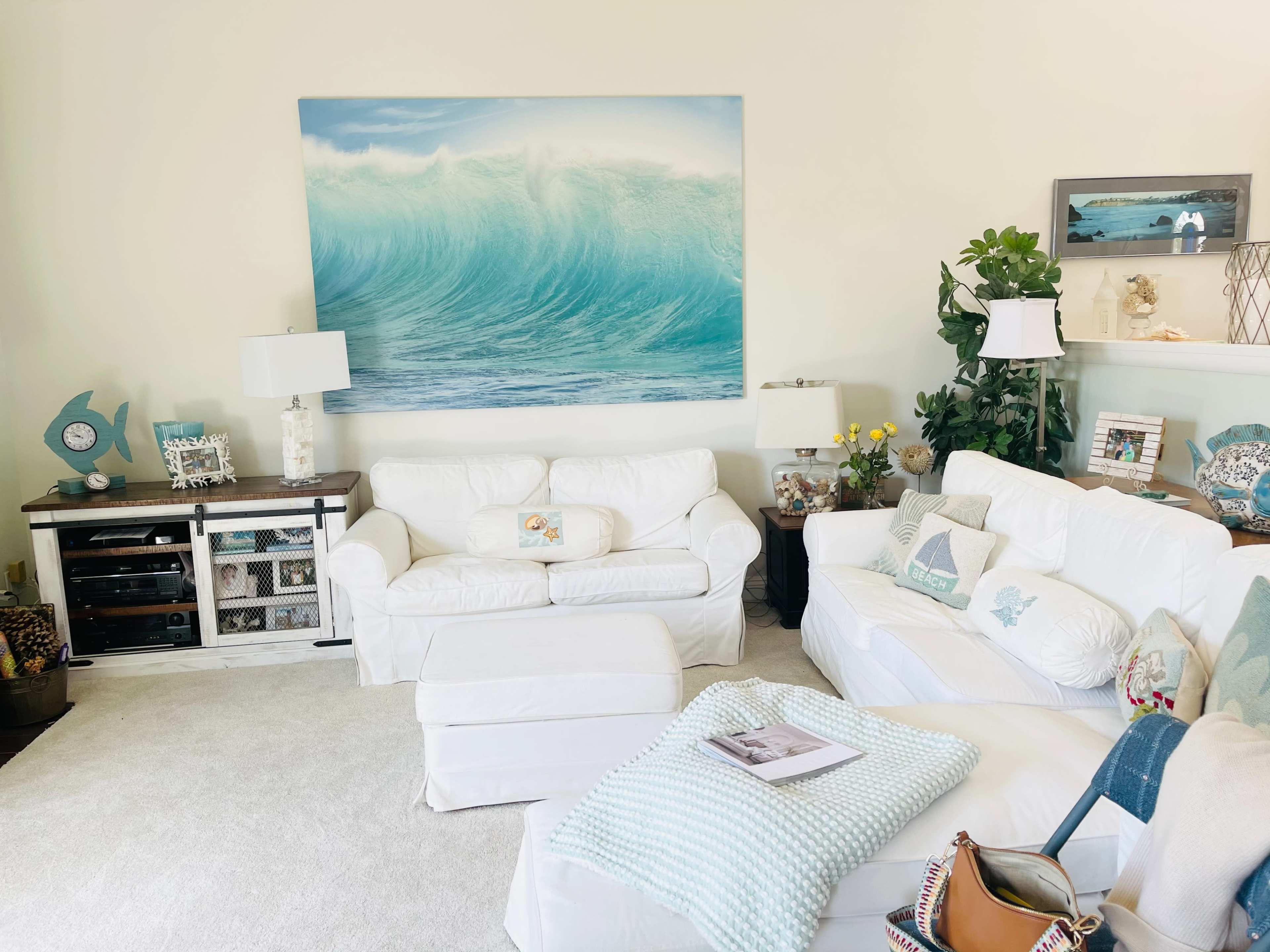 The living room features white furniture arranged around a light carpet, with a large ocean wave painting on the wall and decorative items on shelves.