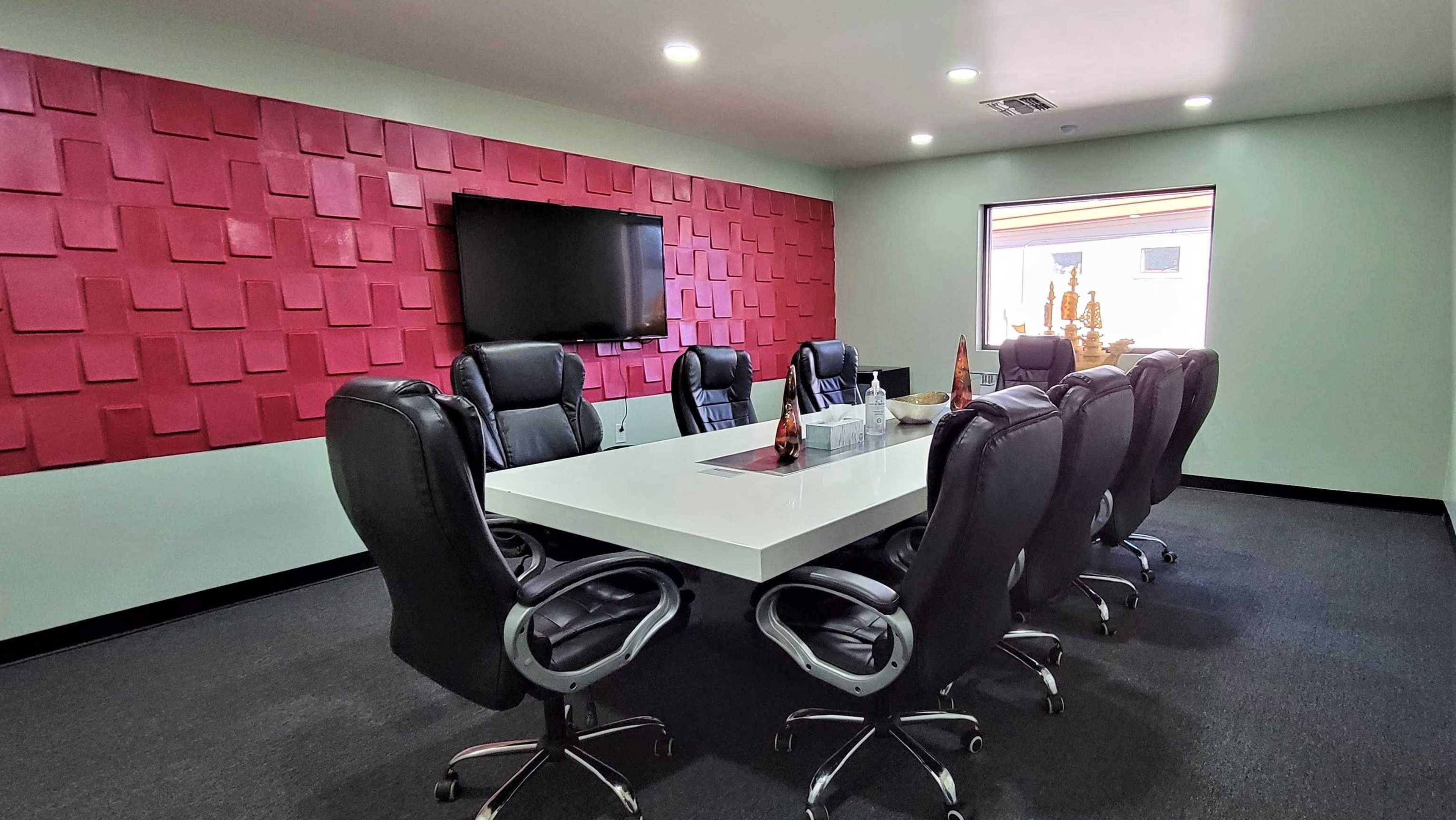 A modern conference room with a large white table surrounded by black ergonomic chairs, a television mounted on the wall, and a textured red accent wall.