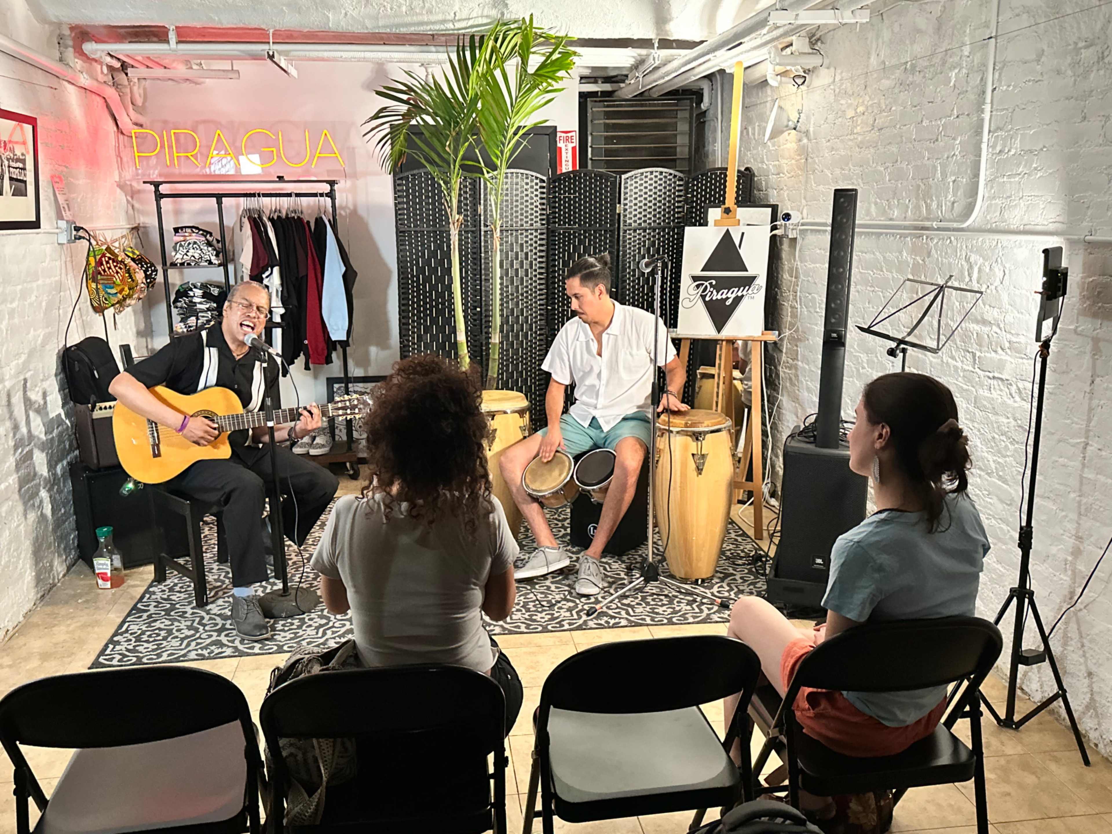 A musician plays guitar while another man drums, performing for an audience seated on black chairs in a small, decorated room.
