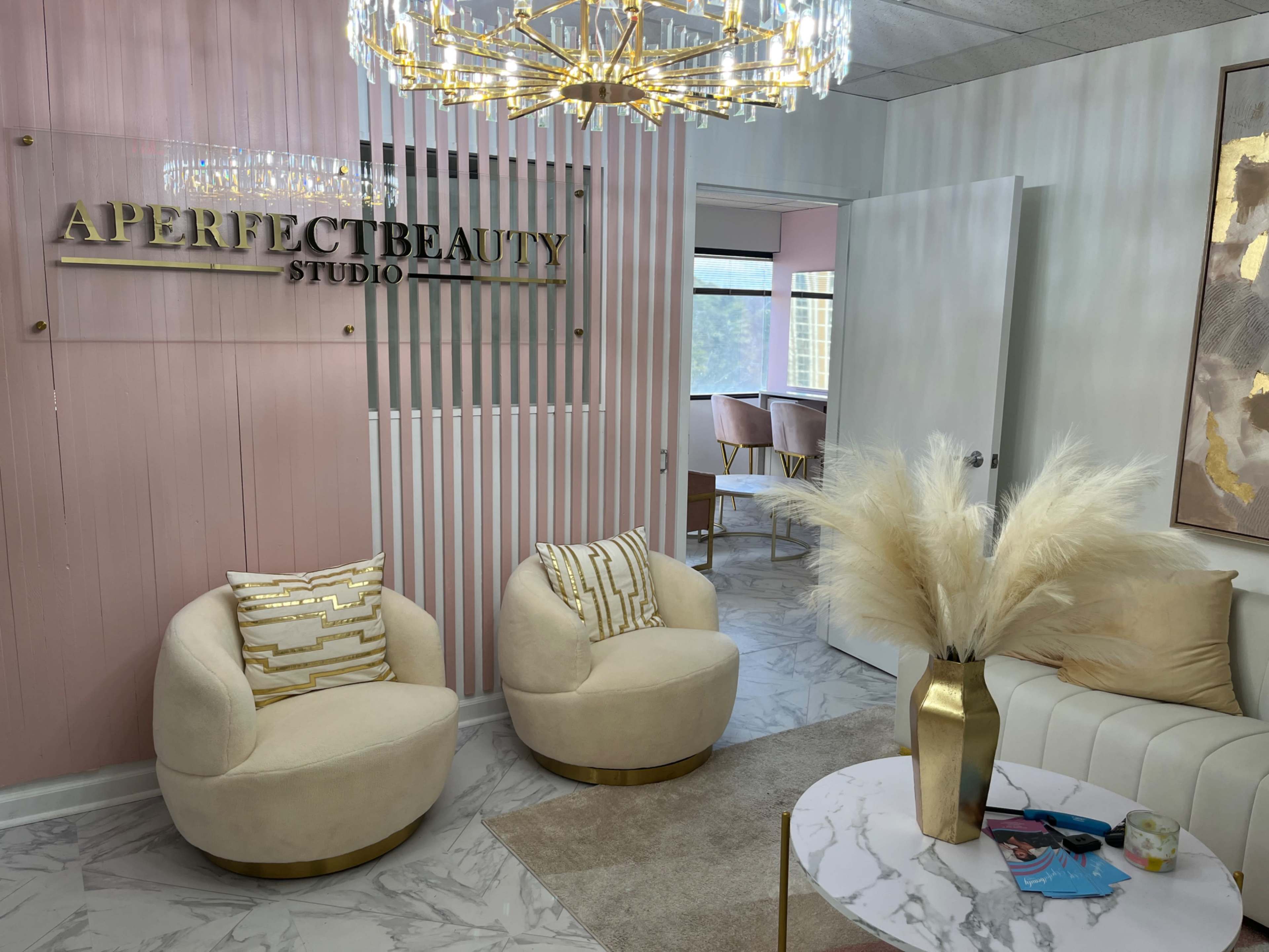 The image shows a modern beauty studio with a light pink wall, a large chandelier, and stylish seating arrangements.
