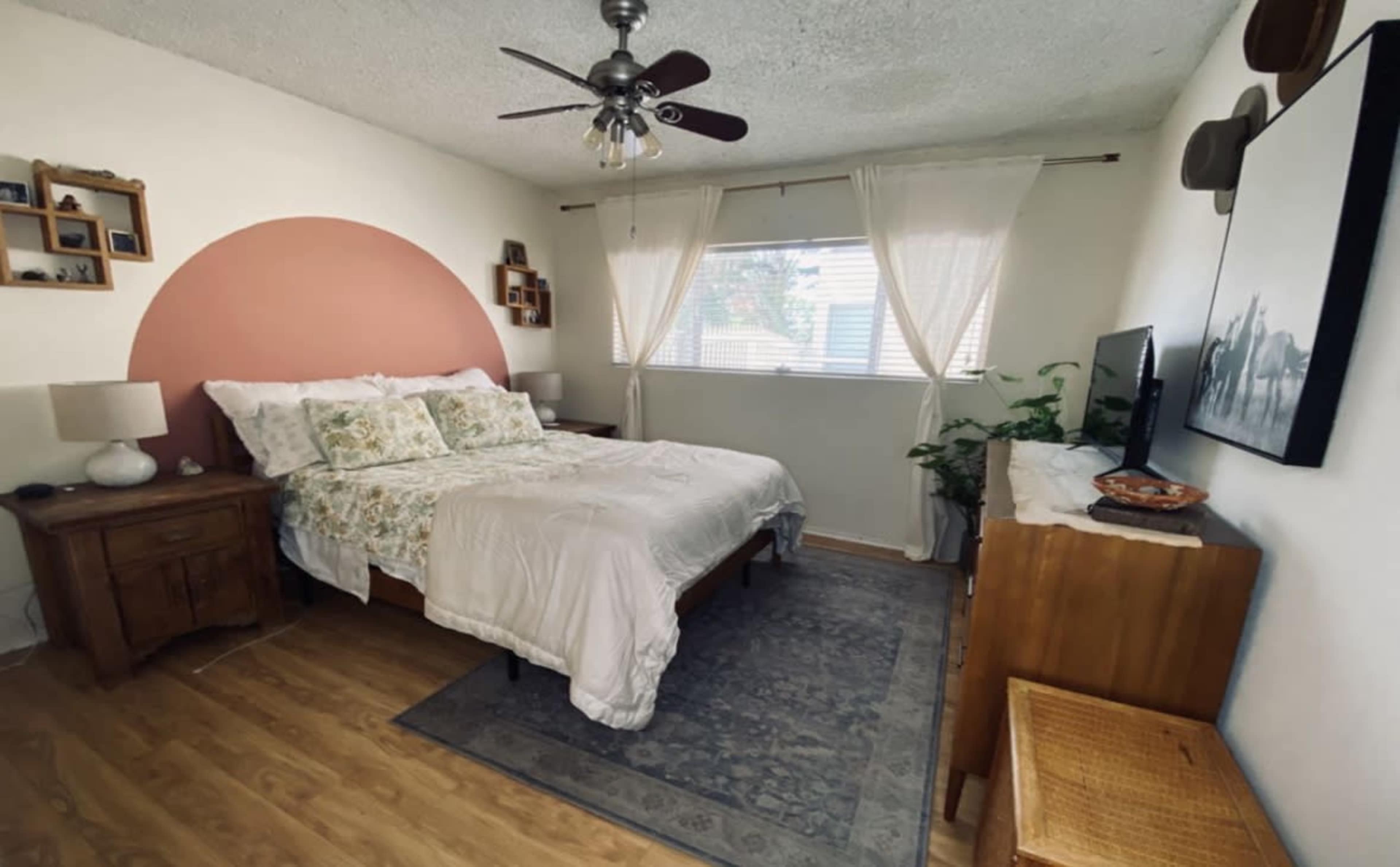 The bedroom features a bed with a light-colored duvet, a wooden nightstand, and a wall adorned with a circular accent in a muted color near a window.