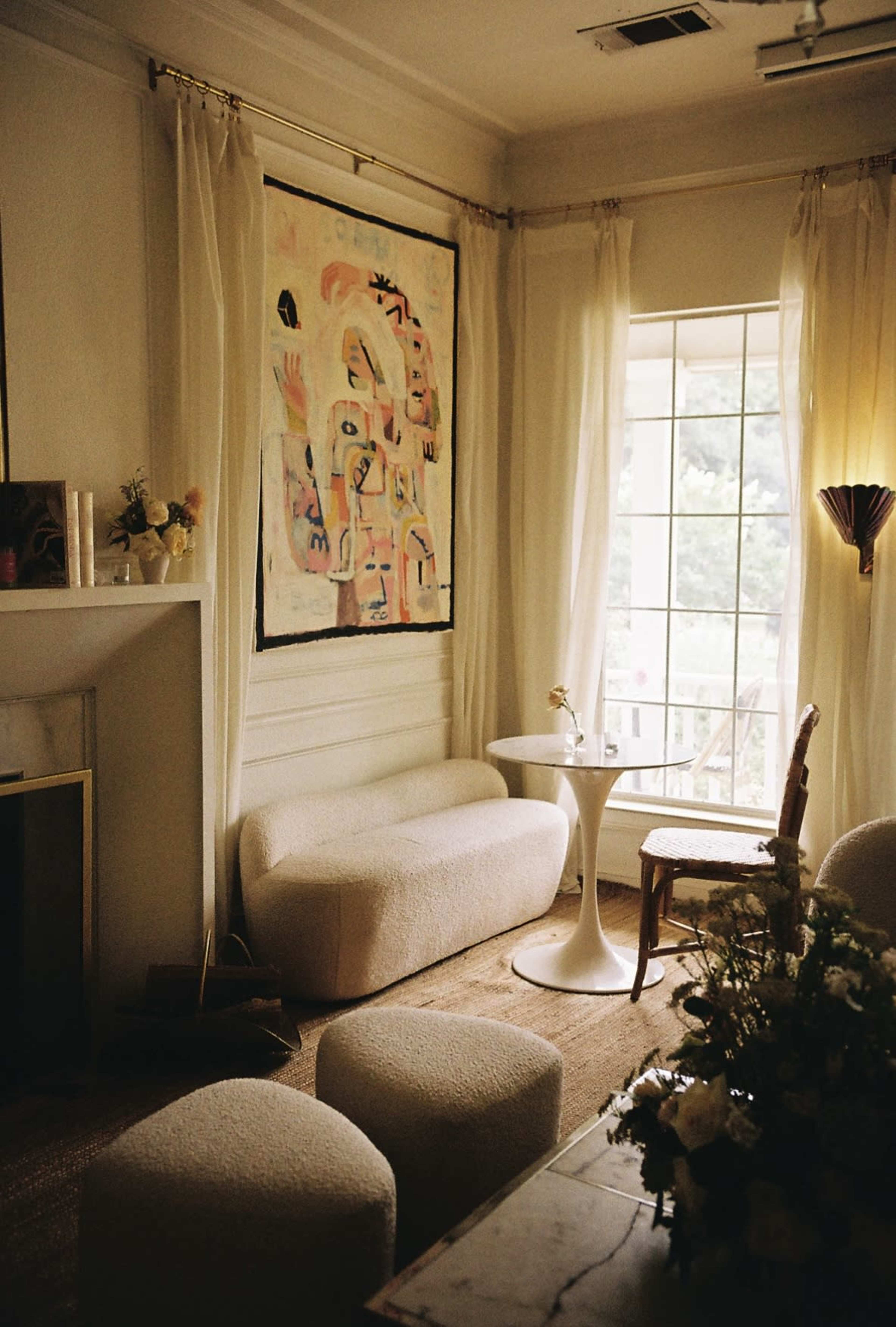 A cozy interior features a sofa and stools, a round table, a chair, and a colorful abstract painting on the wall, illuminated by natural light from a large window.
