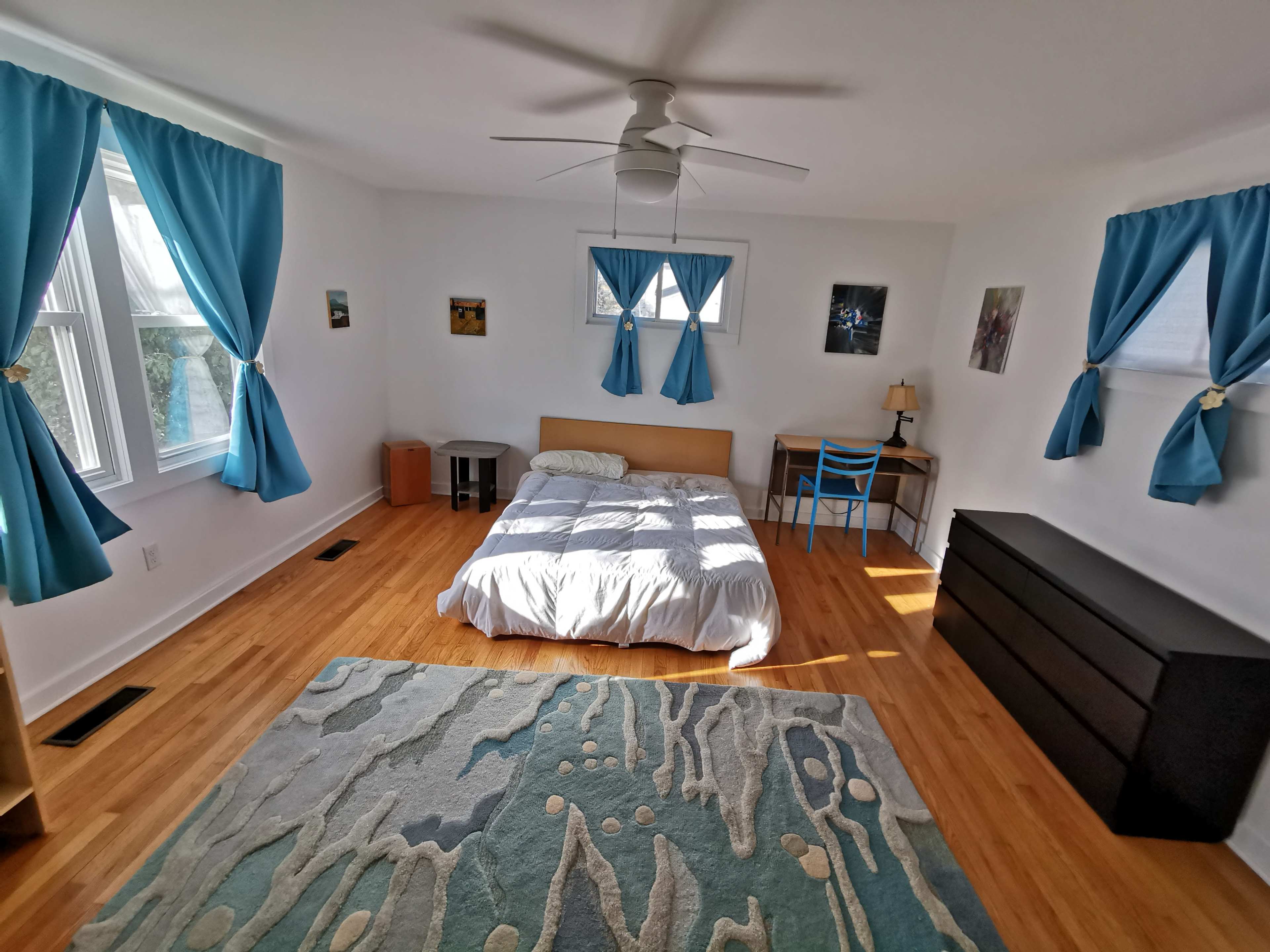A bright bedroom featuring hardwood floors, a bed with white bedding, a desk with a blue chair, and large windows adorned with blue curtains.