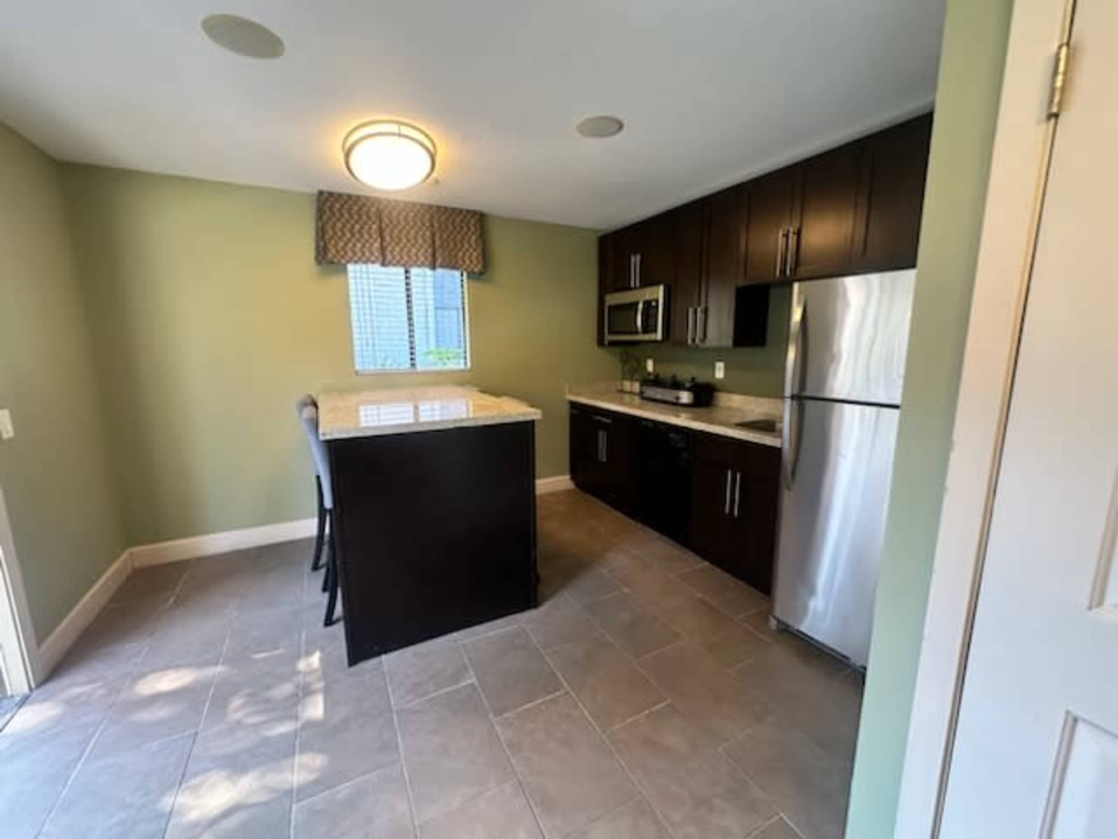 A modern kitchen with dark wood cabinetry, a stainless steel refrigerator, and a small island with two stools, situated under a window with a patterned curtain.