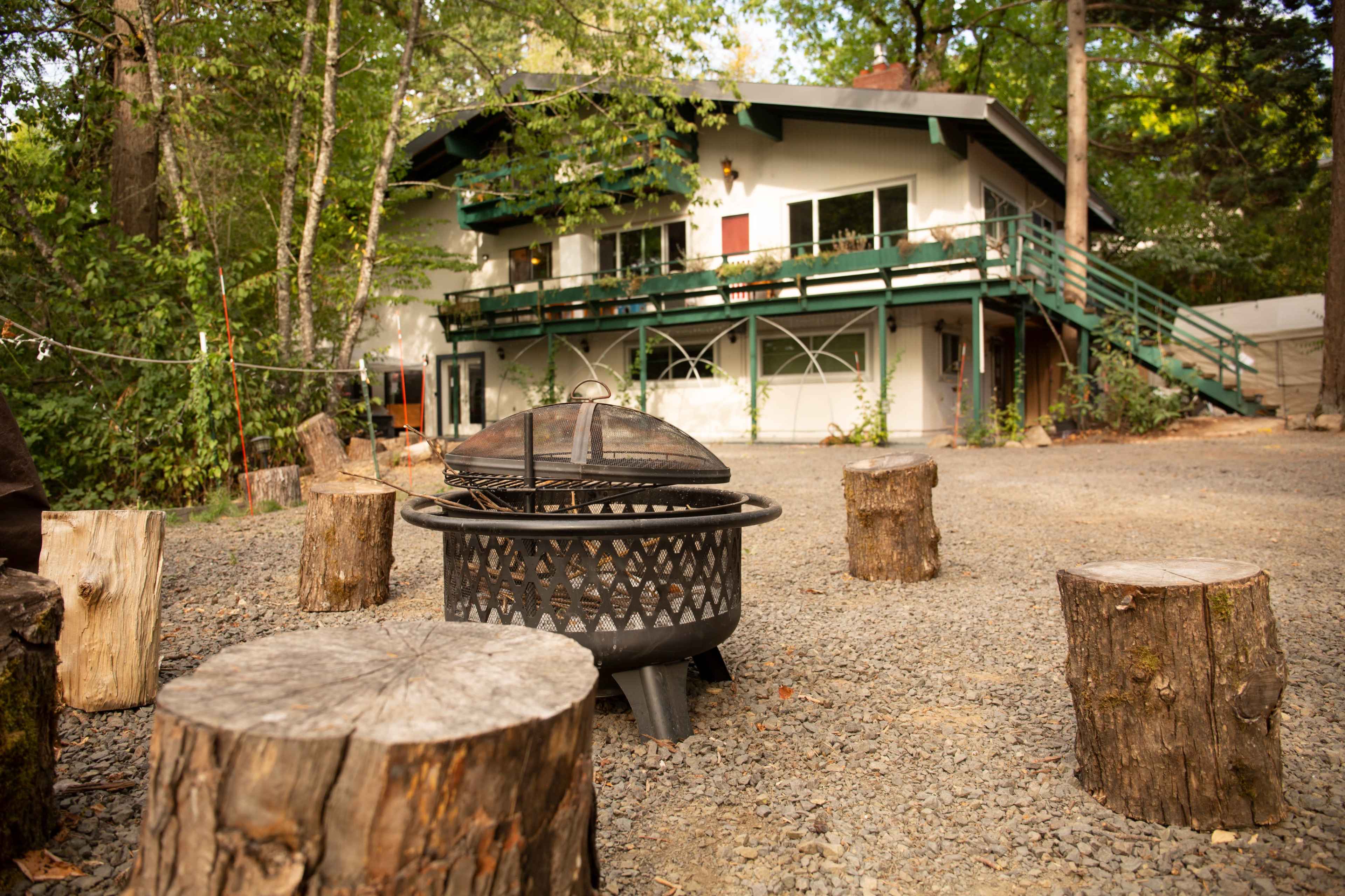 A fire pit sits surrounded by wooden stumps in a gravel area in front of a two-story house nestled among trees.