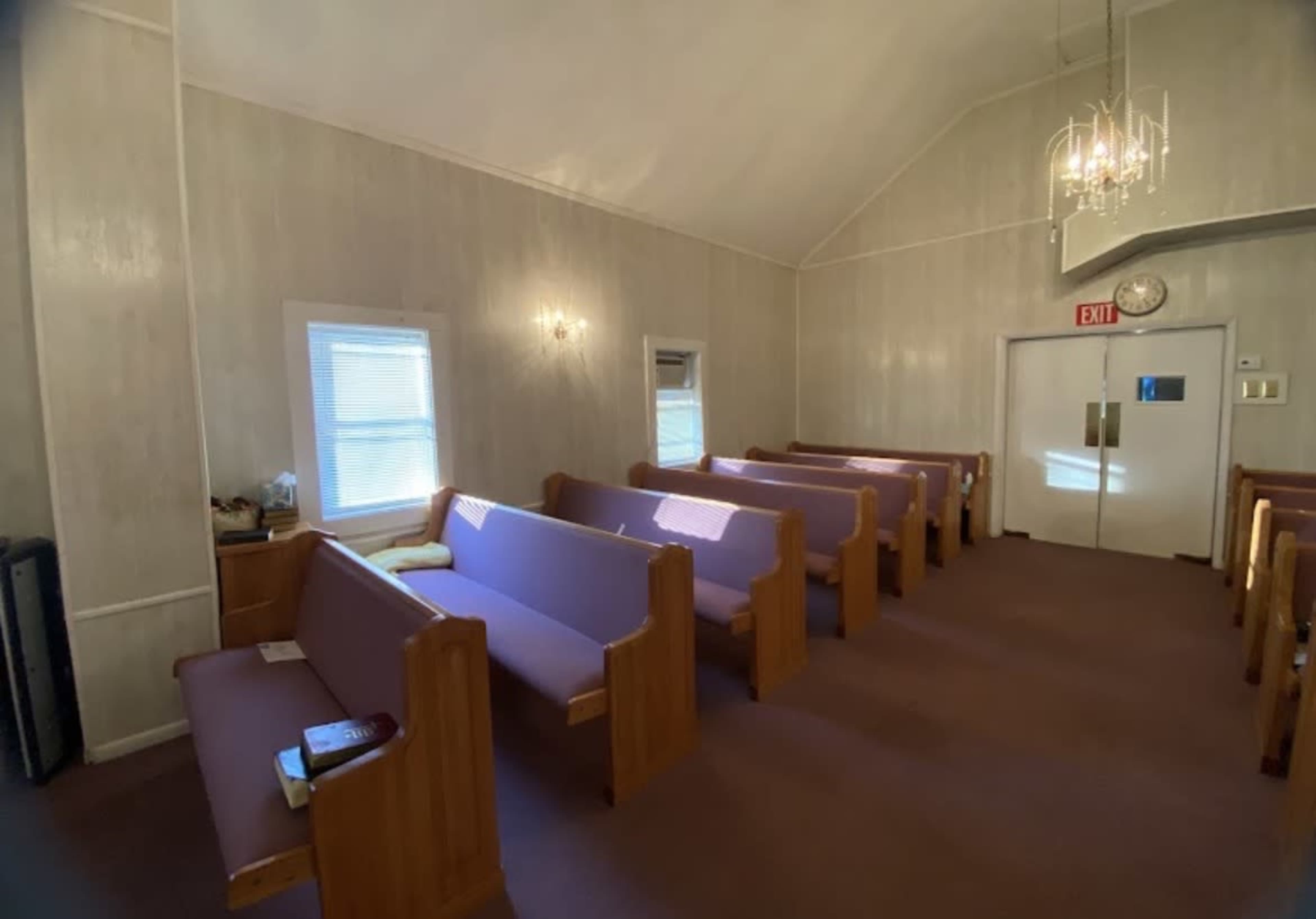 The interior of a small church features several rows of purple pews, with a chandelier hanging from the ceiling and an exit door at one end.