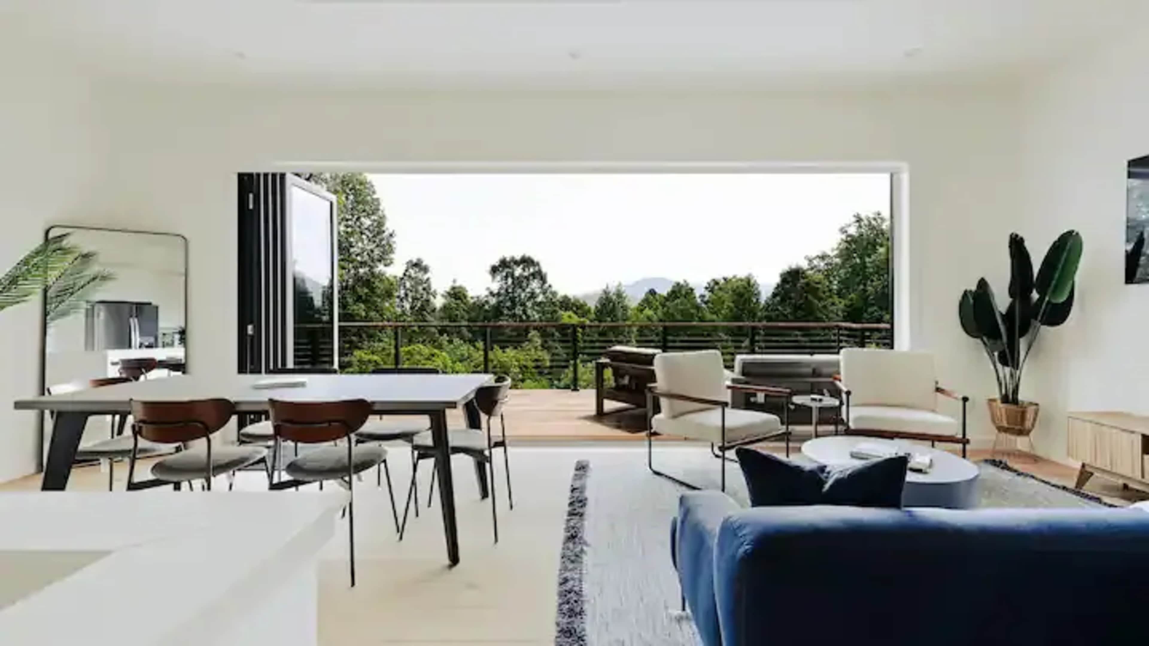 A modern living space with a dining area, a seating area, and large sliding glass doors that open to a deck surrounded by trees.