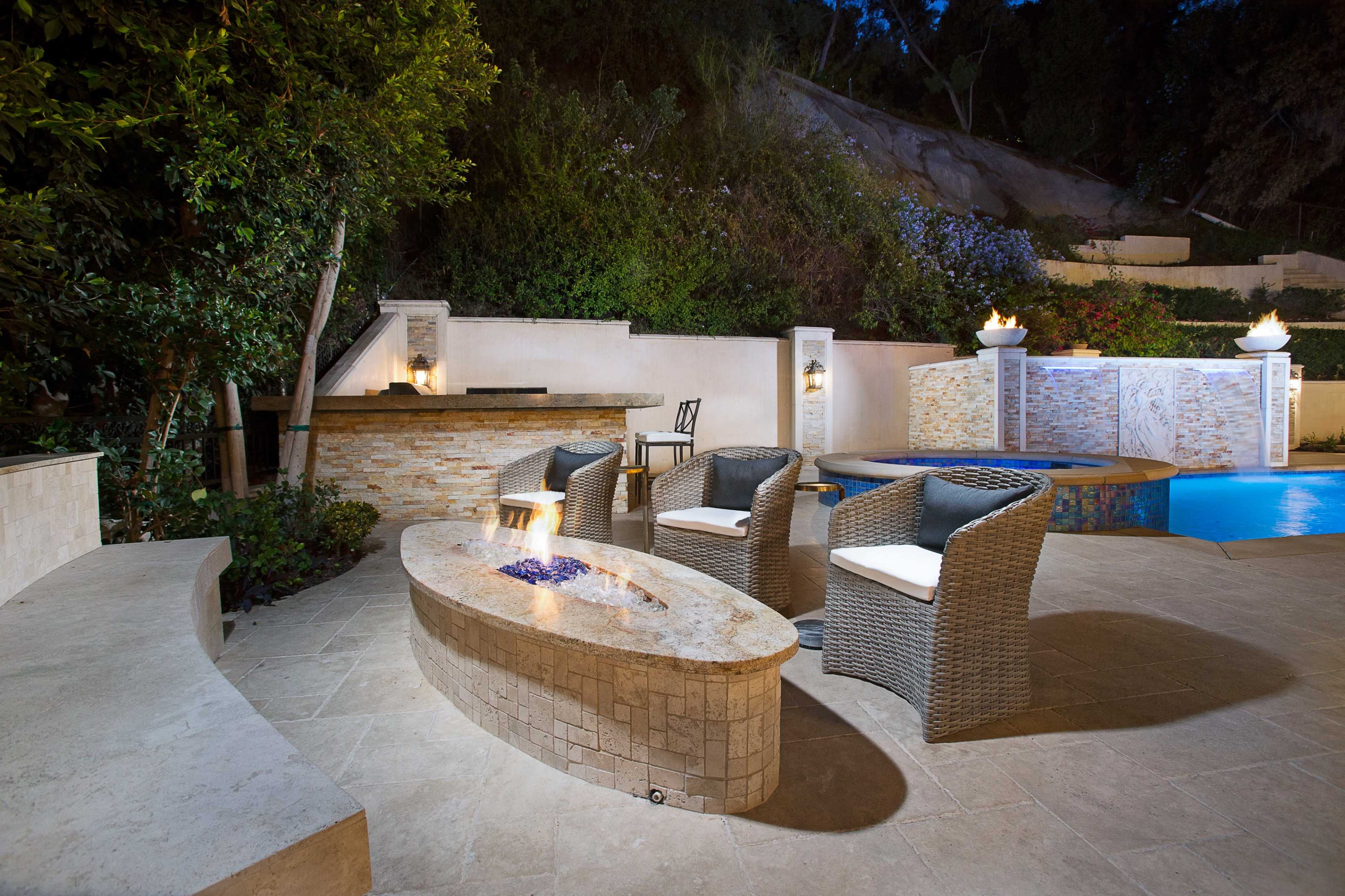 The image shows a modern outdoor patio area featuring a circular stone fire pit surrounded by four wicker chairs, with a swimming pool and landscaped greenery in the background.