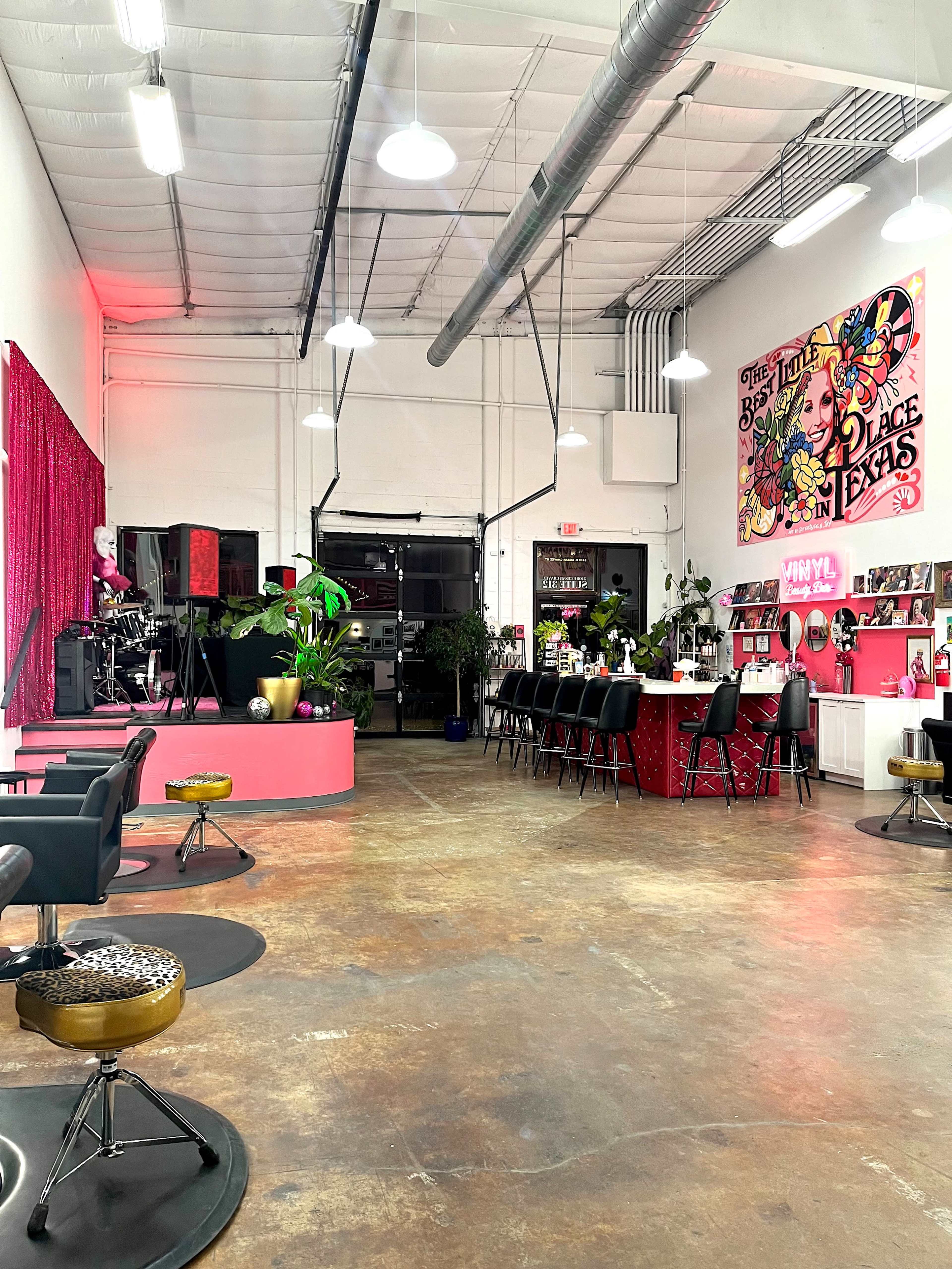The image shows a brightly lit hair salon featuring a mix of pink and black furniture, with a reception area and styling stations arranged against the white walls.