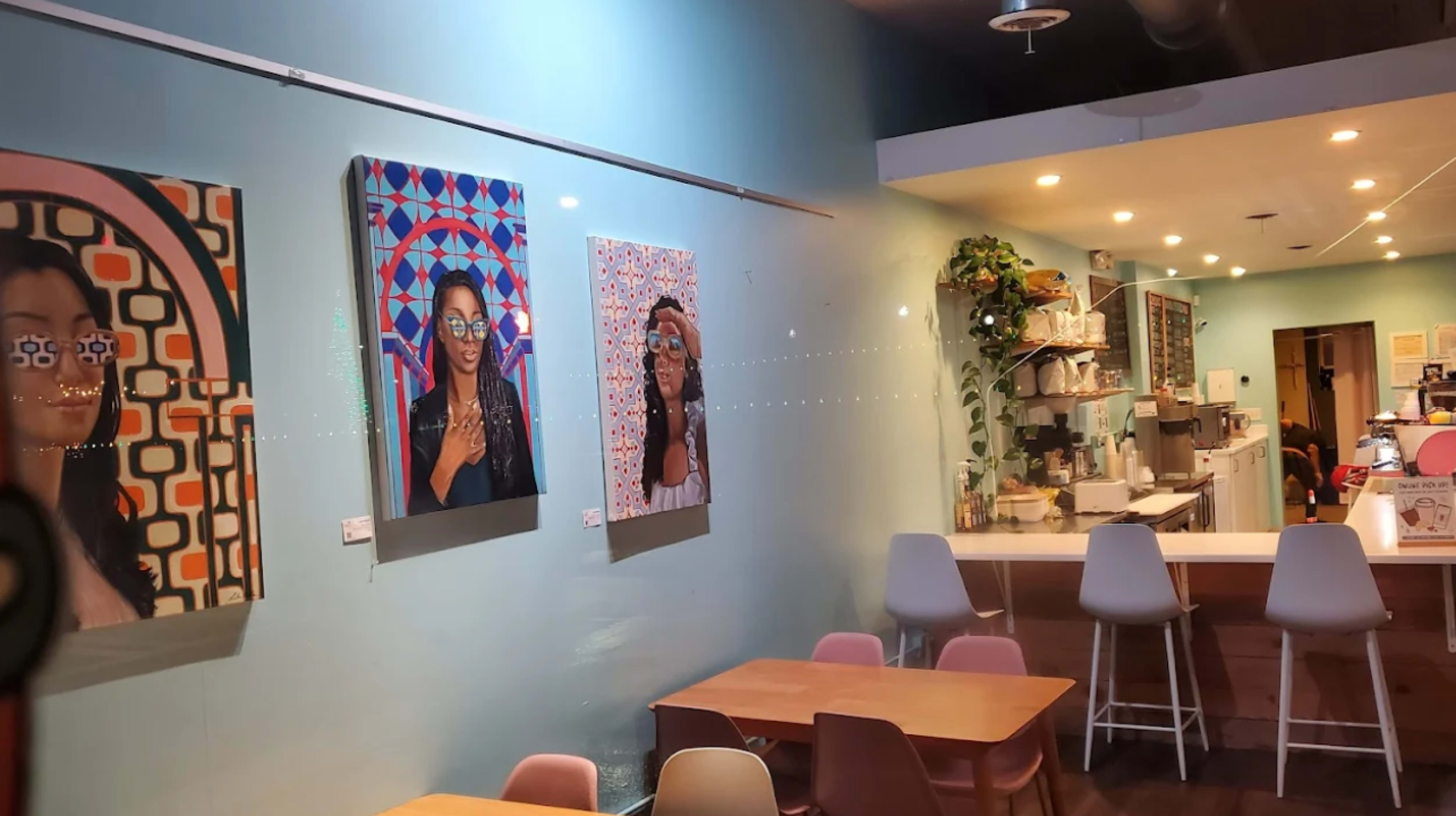 The image shows an indoor café featuring colorful artwork on the walls, a counter with food and drinks, and wooden tables arranged for customers.