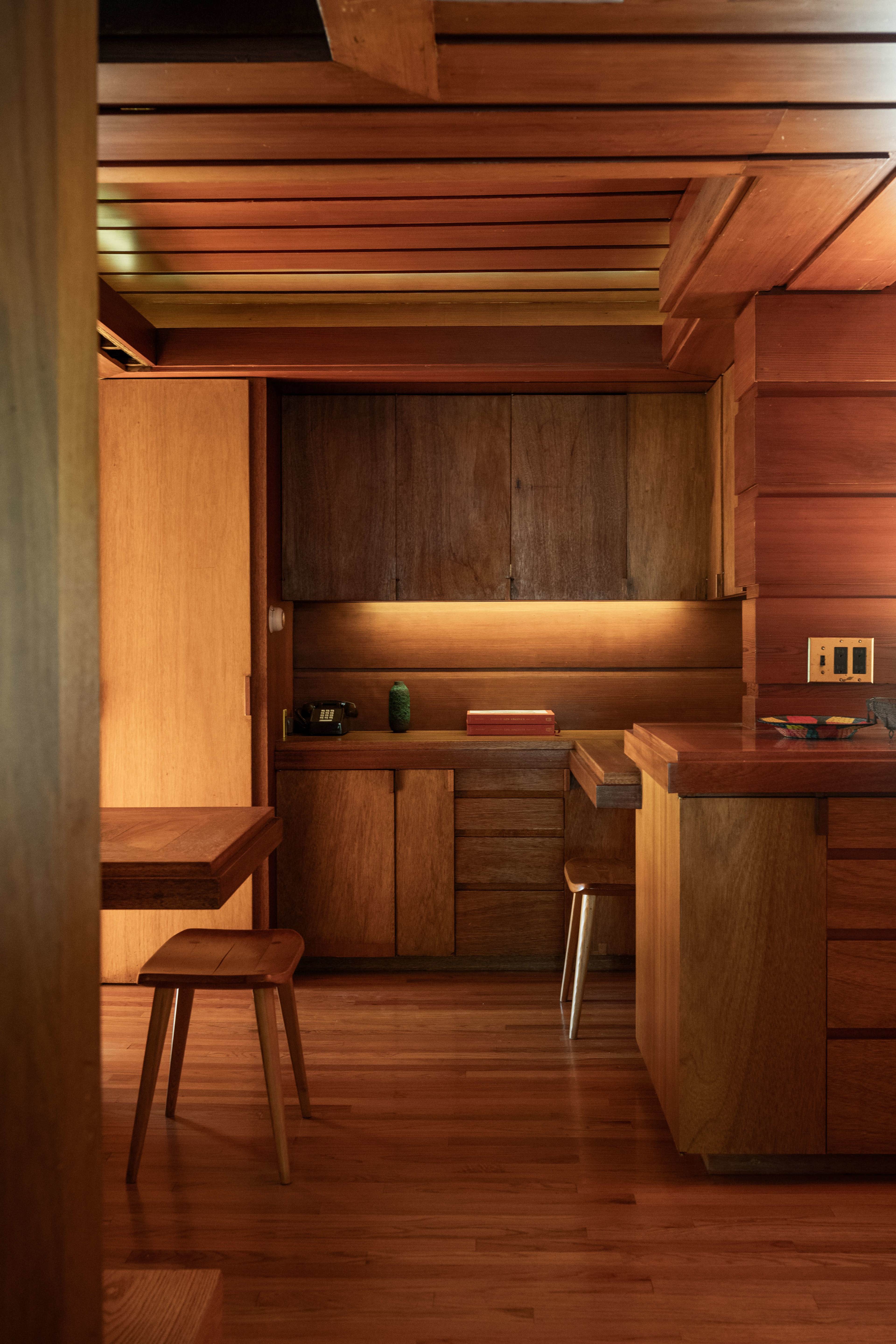 A wooden interior of a modern kitchen with a table, stools, and low cabinetry under warm lighting.