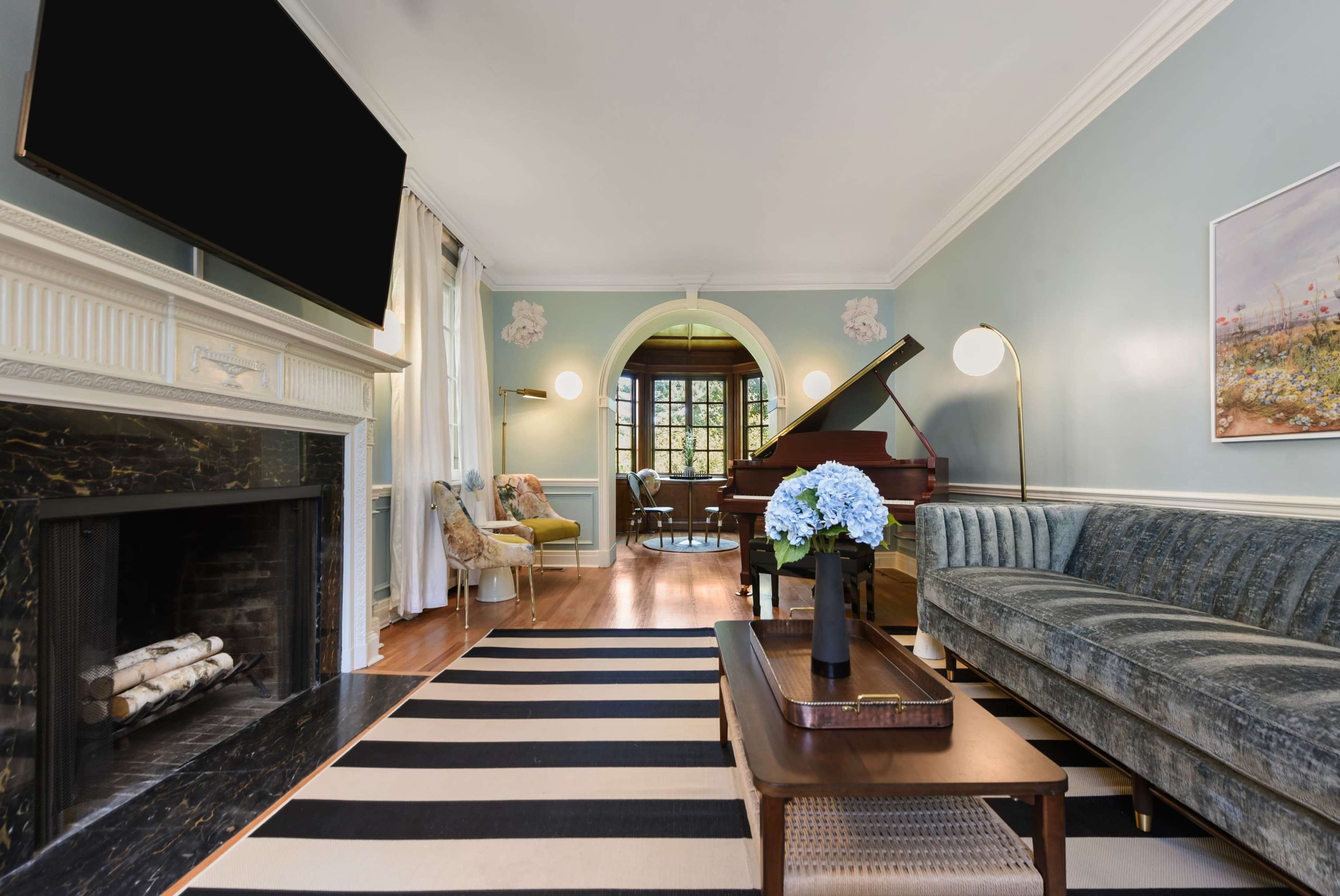 The living room features a grand piano, a fireplace, and a sofa facing a striped rug under a coffee table with a vase of flowers.