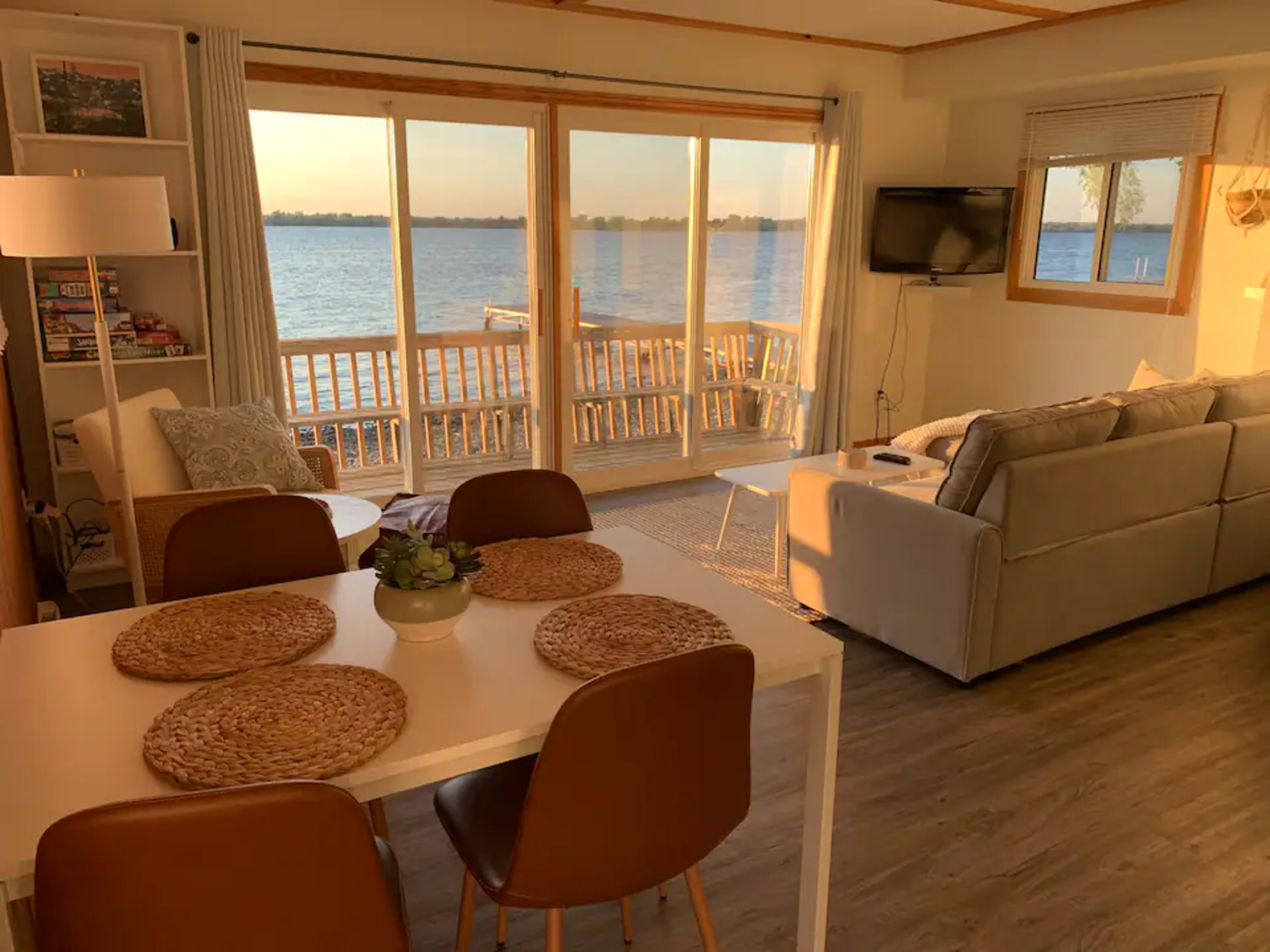 The image shows a cozy living space with a dining area, a couch, and large sliding glass doors overlooking a waterfront view.