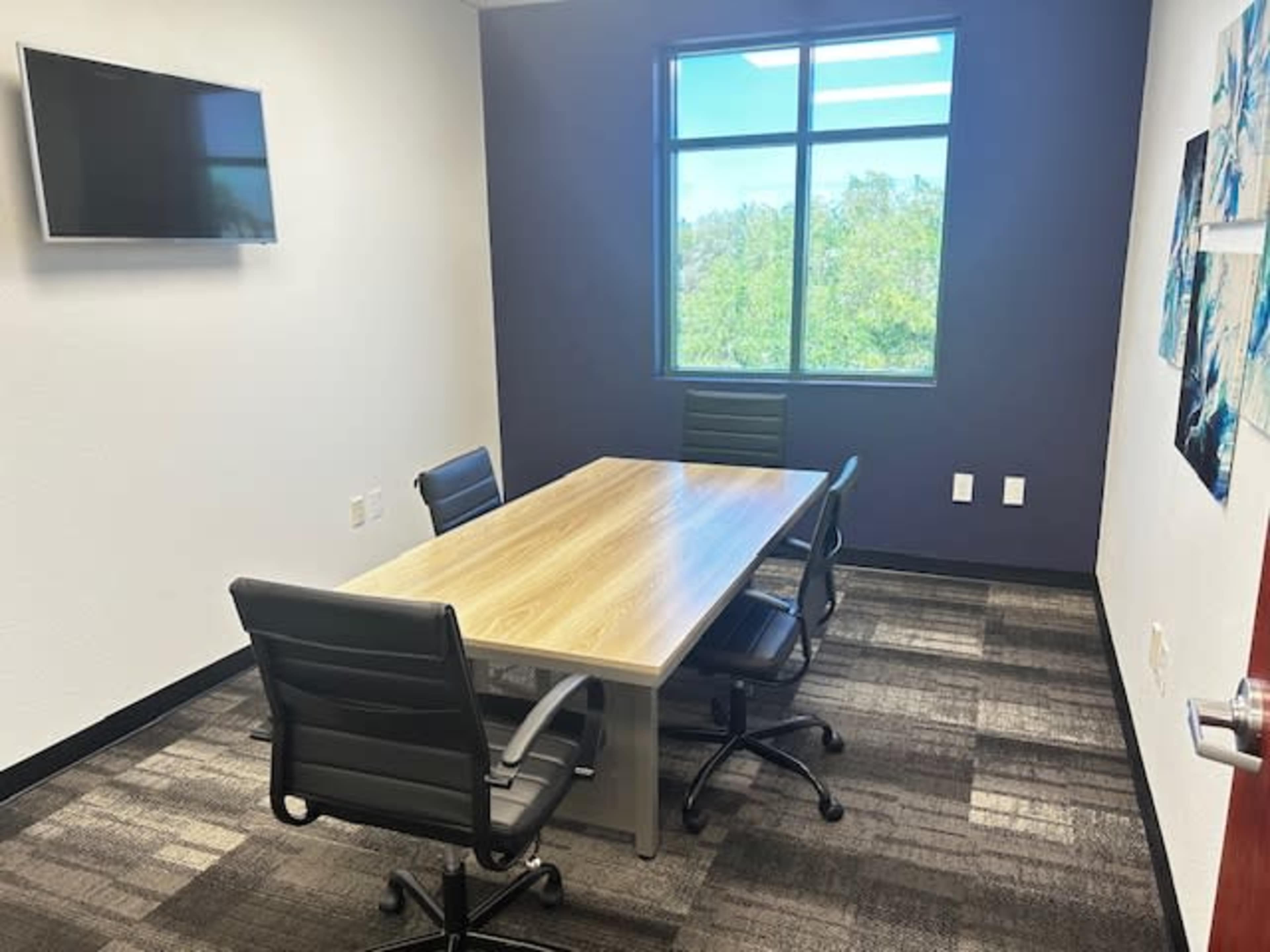 A conference room features a wooden table surrounded by four black chairs, with a wall-mounted TV and large window offering a view of greenery outside.