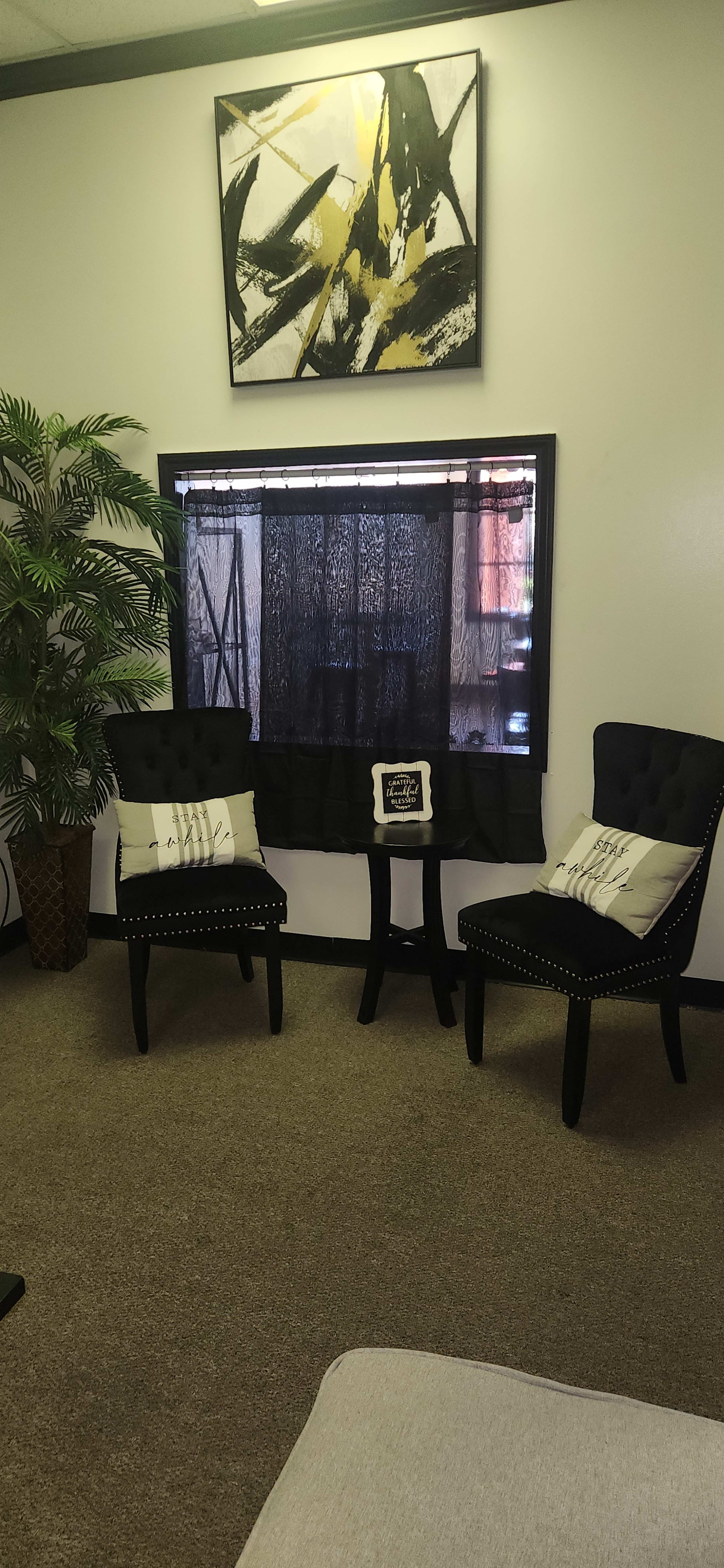 The image shows a small seating area with two black chairs, a decorative plant, and a window covered by sheer curtains, all set against a neutral wall.