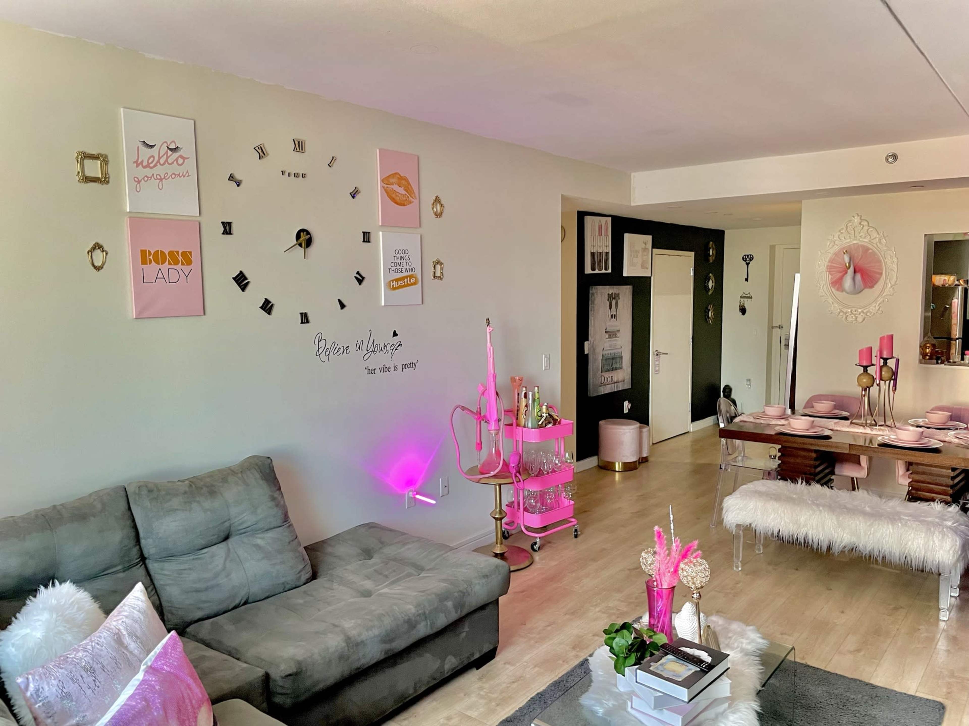 The room features a modern living area with a gray couch, a pink-themed dining table, and various decorative items on the walls.