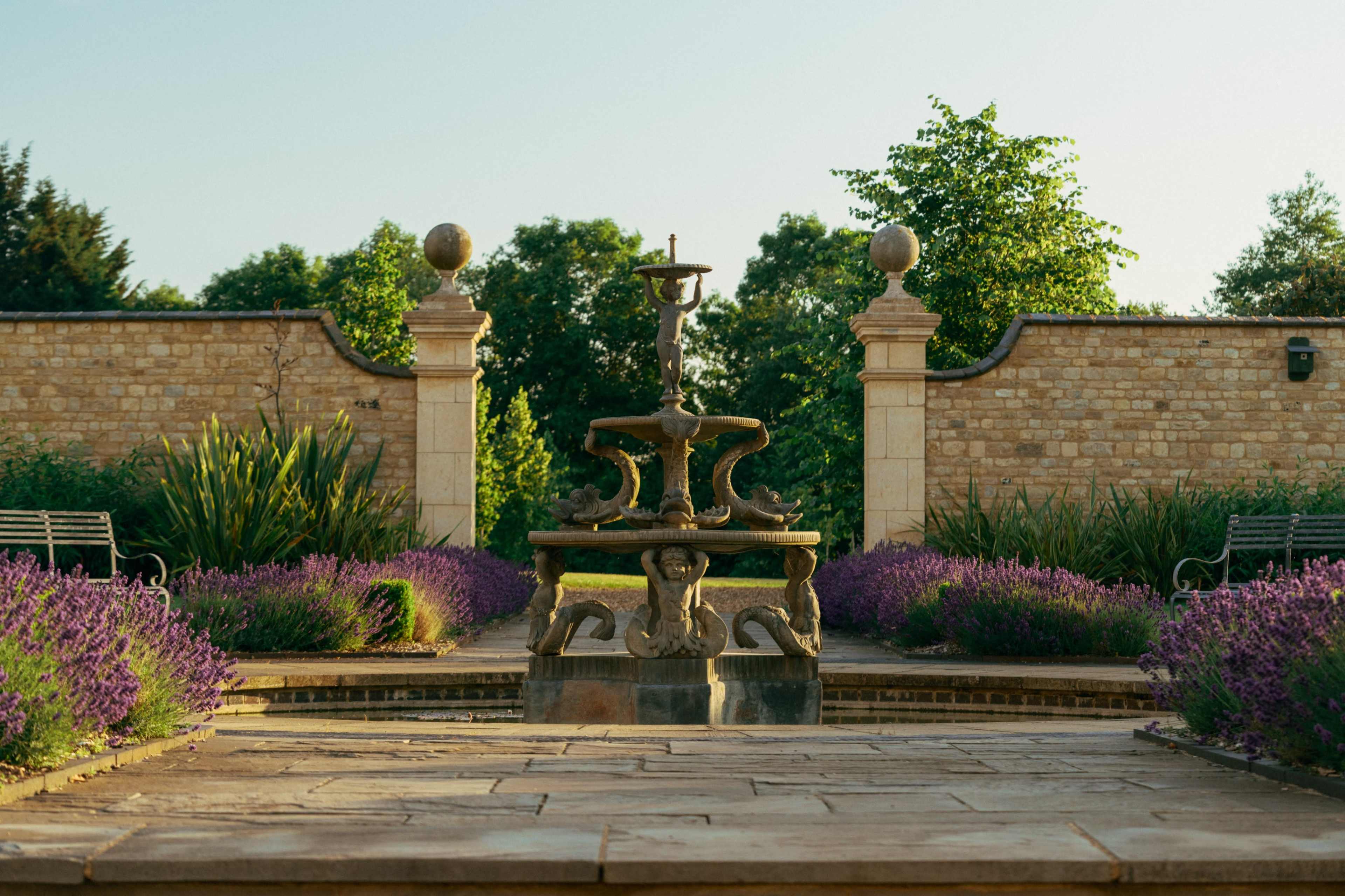 The image shows a stone fountain surrounded by neatly trimmed lavender bushes in a well-maintained garden with a stone wall and ornamental trees in the background.