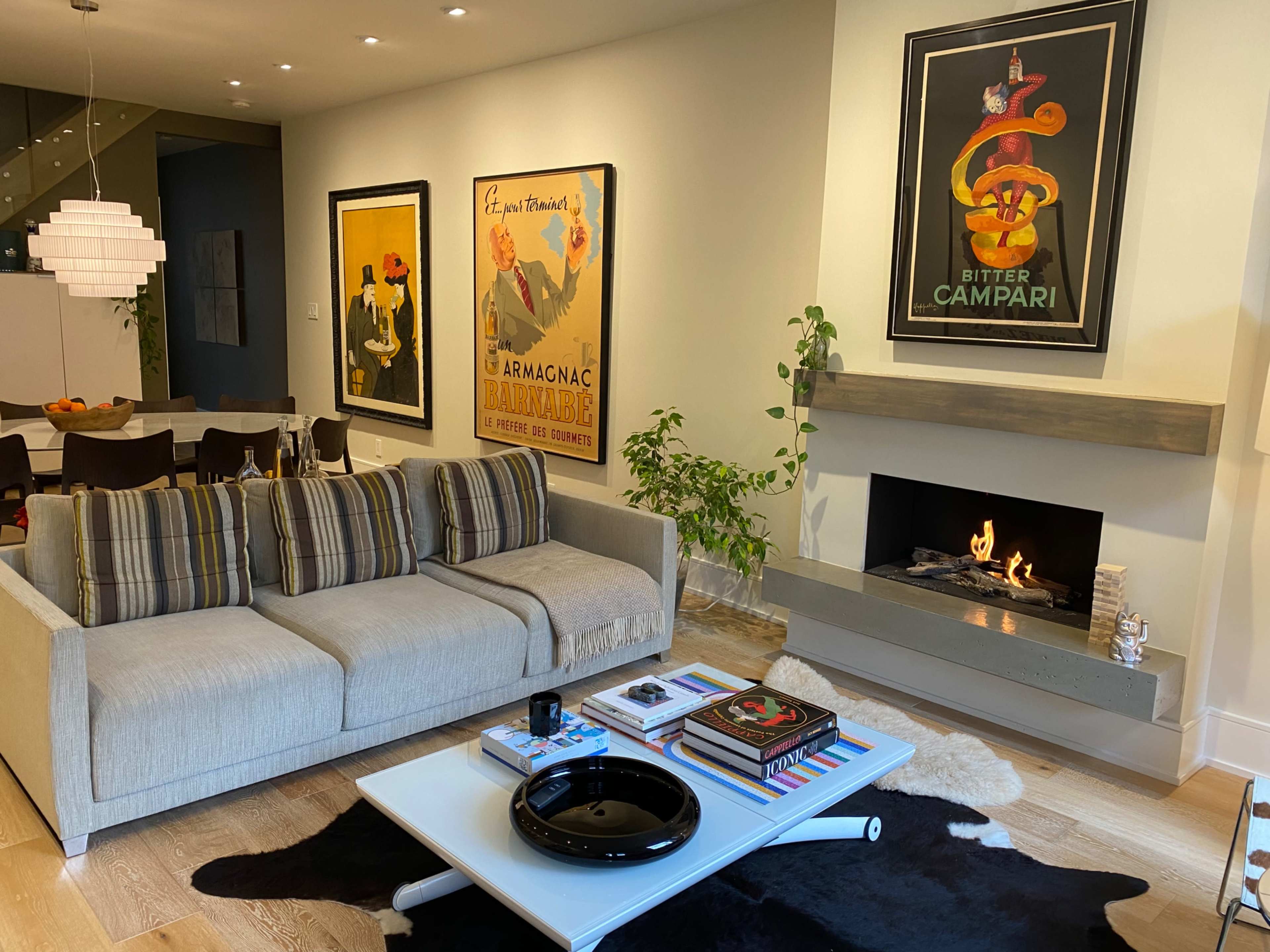 A modern living room with a light gray sofa, two framed vintage posters on the walls, a black coffee table with books and a decorative dish, and a fireplace with a visible flame.