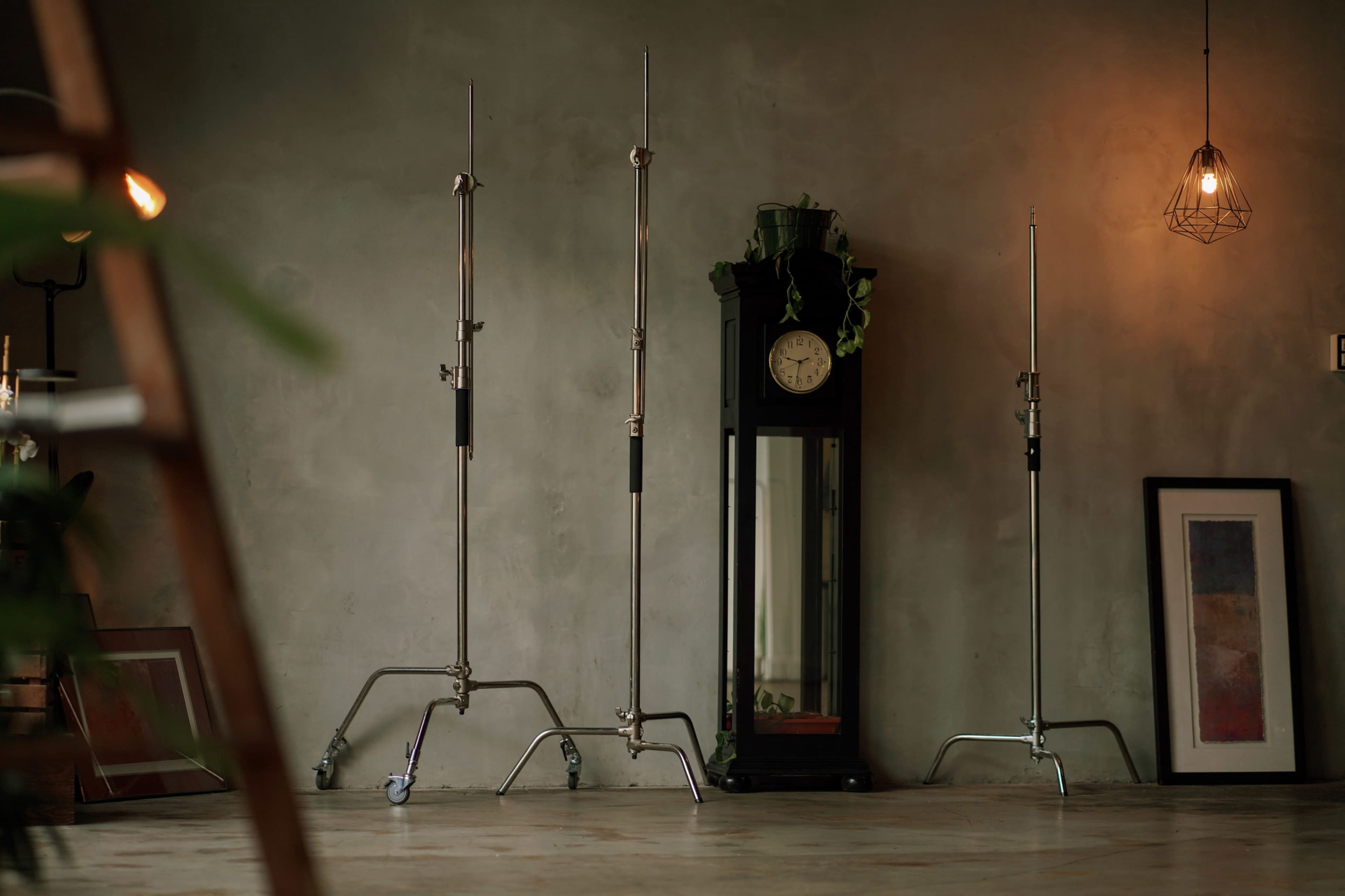The image shows a minimalist interior space with three metal light stands positioned near a tall, dark clock tower against a concrete wall, accompanied by a framed artwork and a single pendant light overhead.