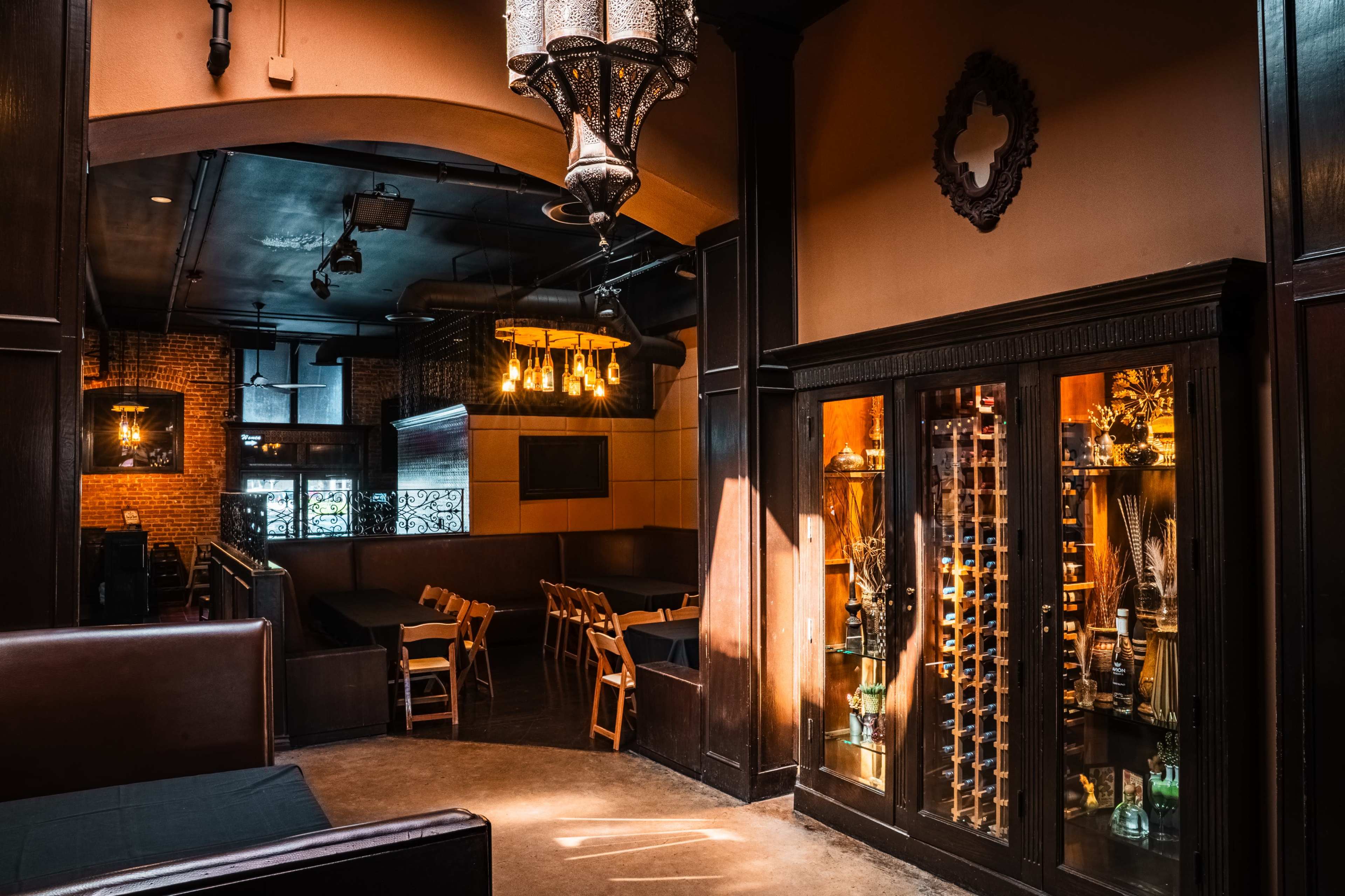 The image shows a dimly lit interior of a restaurant or bar, featuring a wooden display cabinet and a seating area with wooden chairs.