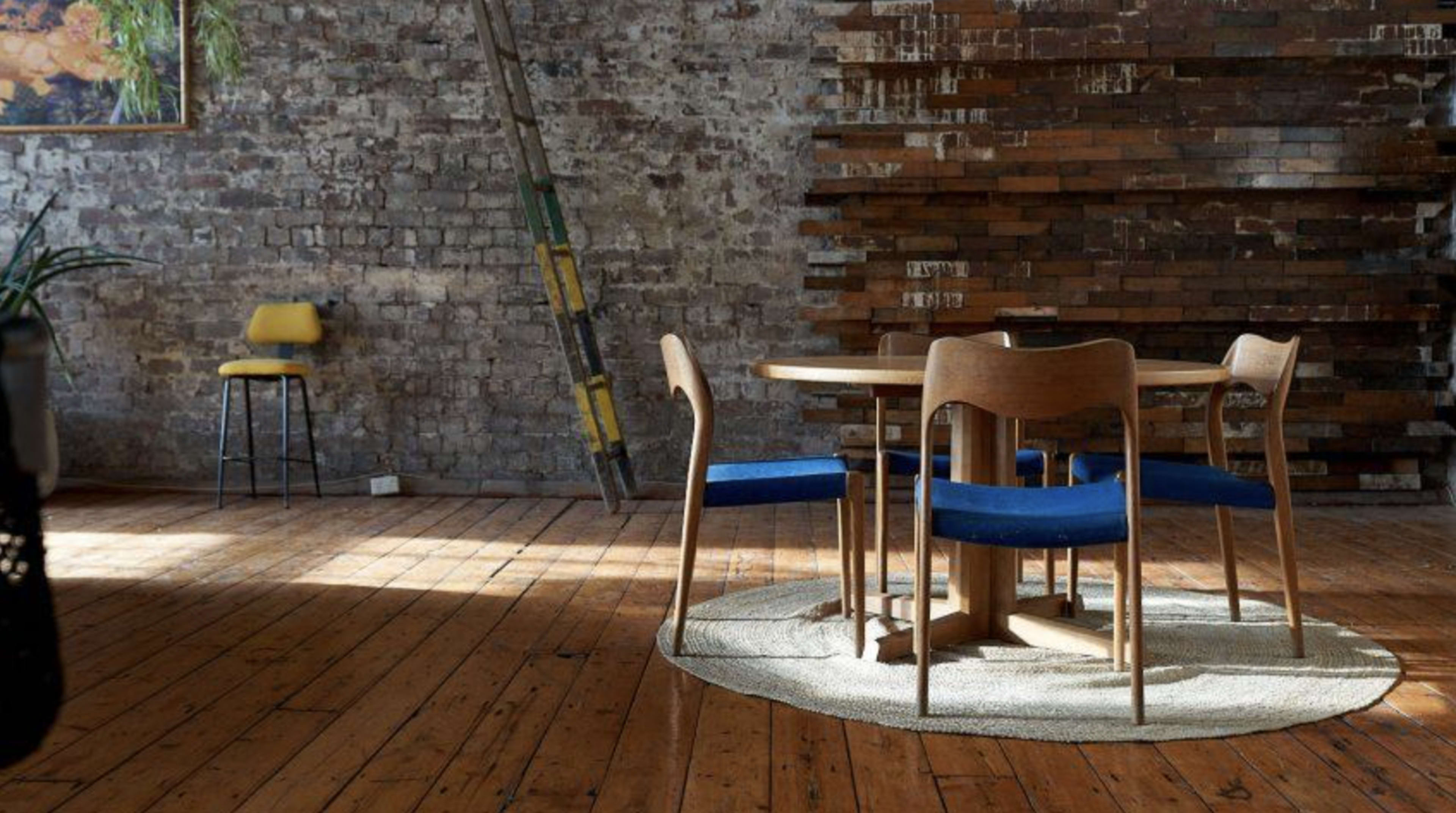 A round wooden table with blue chairs sits on a circular rug in a room with exposed brick walls and wooden flooring.