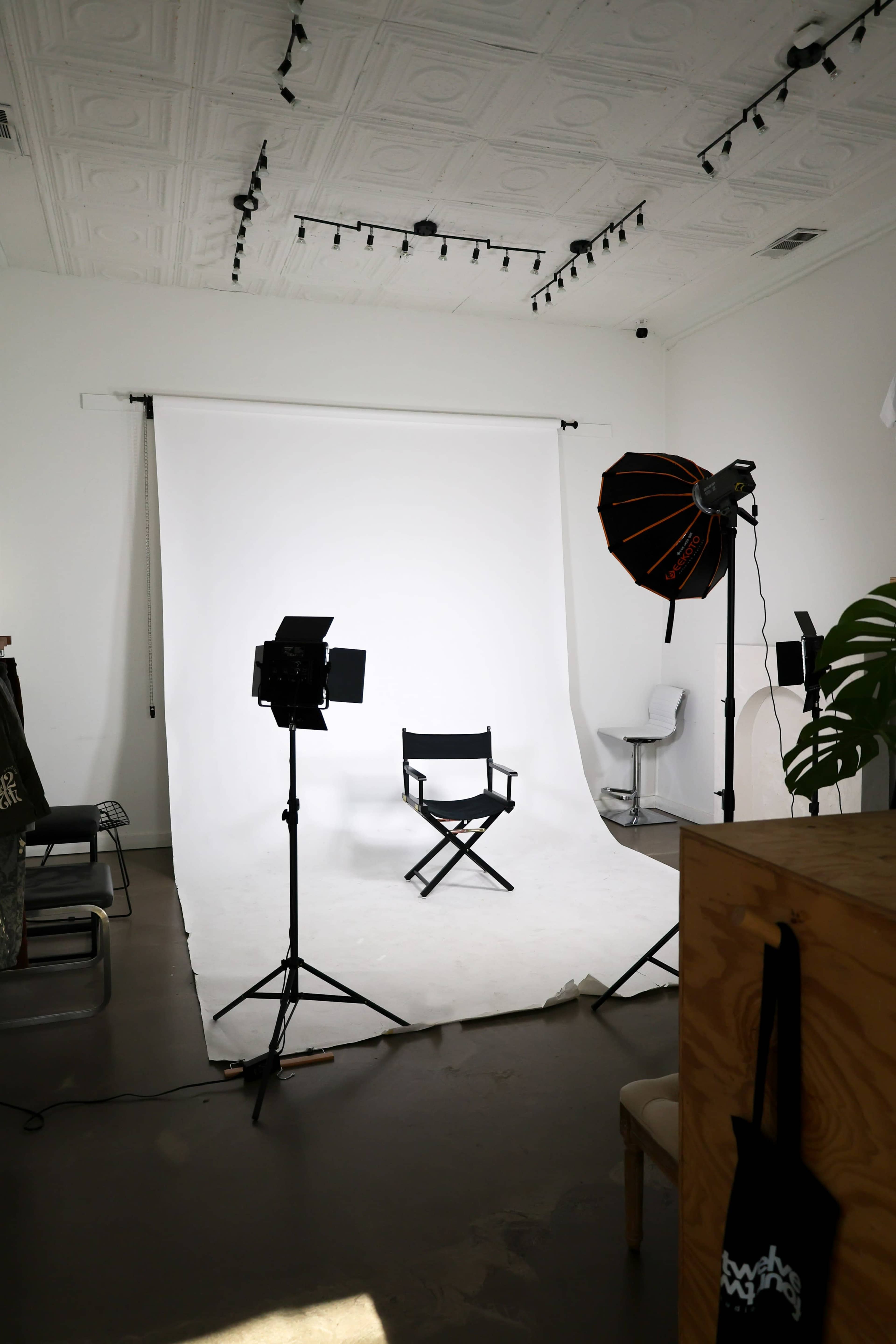 The image shows a photography studio featuring a white backdrop, a black director's chair, and lighting equipment set up around the space.
