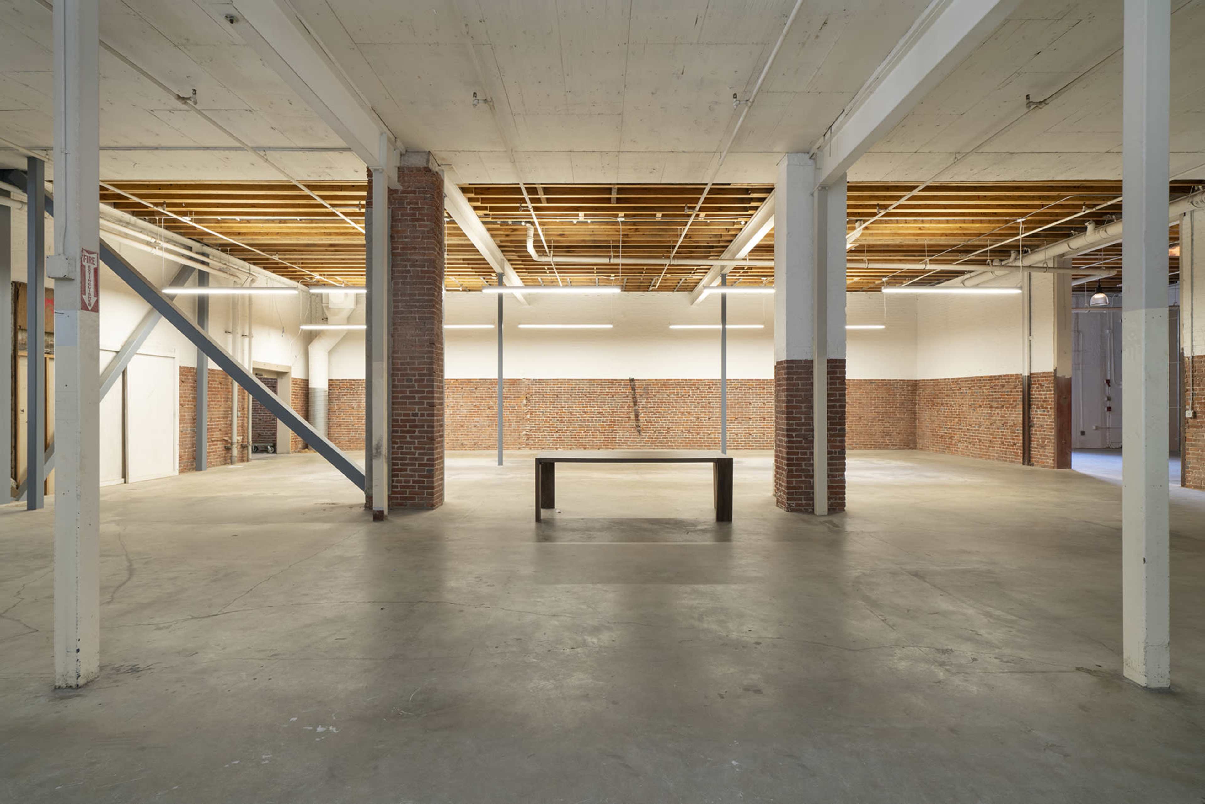 A spacious, empty industrial interior with concrete floors, exposed brick walls, and a central table supported by metal beams.