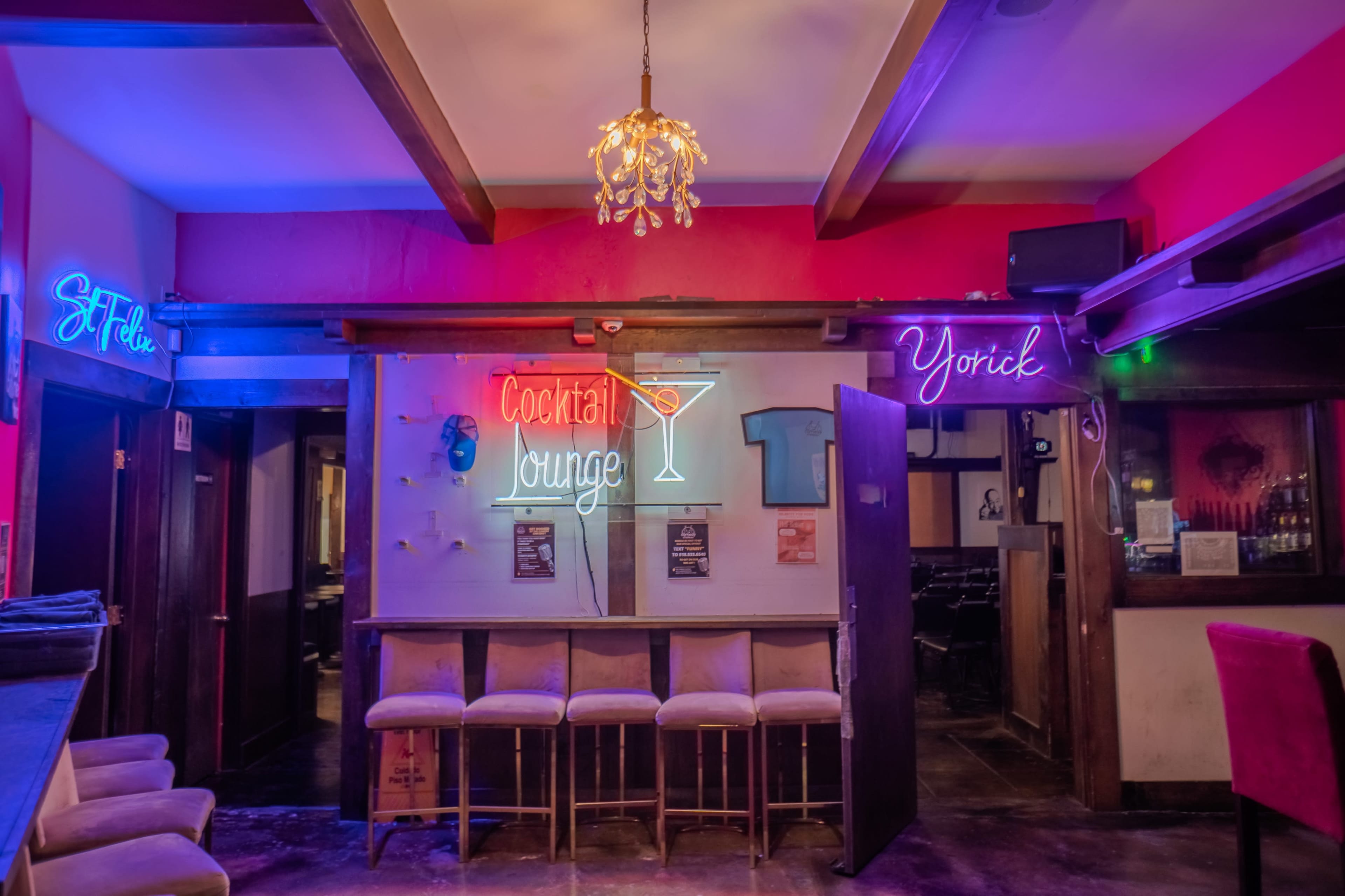 A dimly lit bar area with neon signs labeled "Cocktail Lounge," "Yorick," and "St. Tide" amidst empty stools.